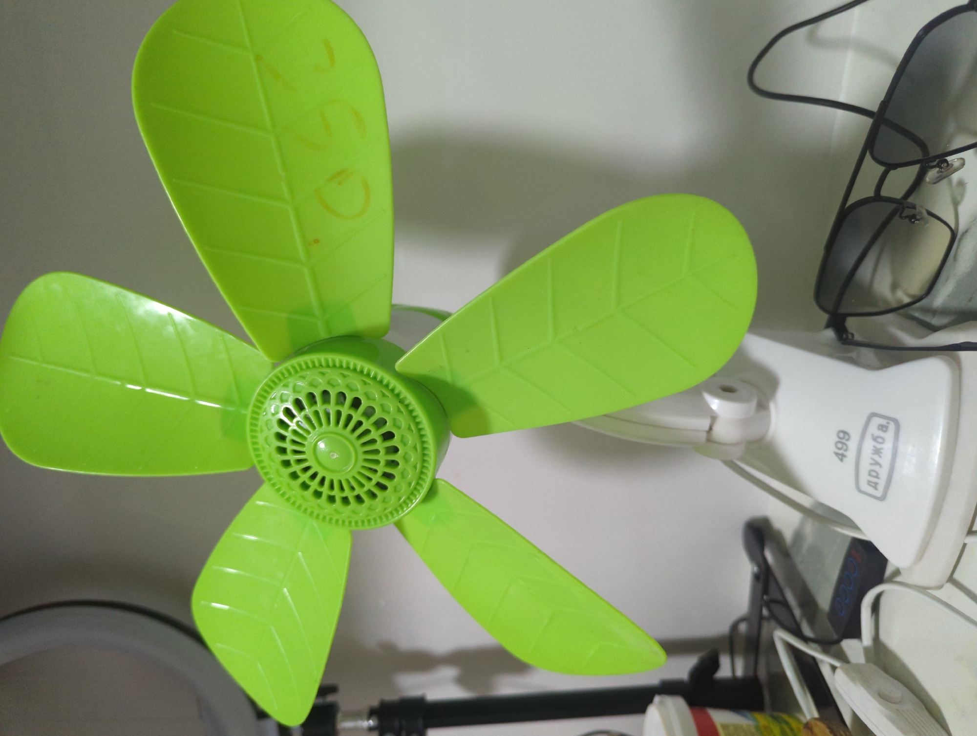 A green electric fan with leaf-shaped blades is positioned in the foreground, angled slightly to the right. The fan's base is white and features the text "499" and "ДРУЖБА" in grey lettering. To the right of the fan, a pair of black sunglasses with gradient lenses is visible. In the background, a ring light is partially visible on the left, and a white wall serves as the backdrop. The scene appears to be indoors, likely on a surface like a table or shelf, with some electrical cords and a container to the left, suggesting a personal workspace or living area. The location of the image is identified as a village in Kyrgyzstan, specifically, the village of Üç-Döbö.