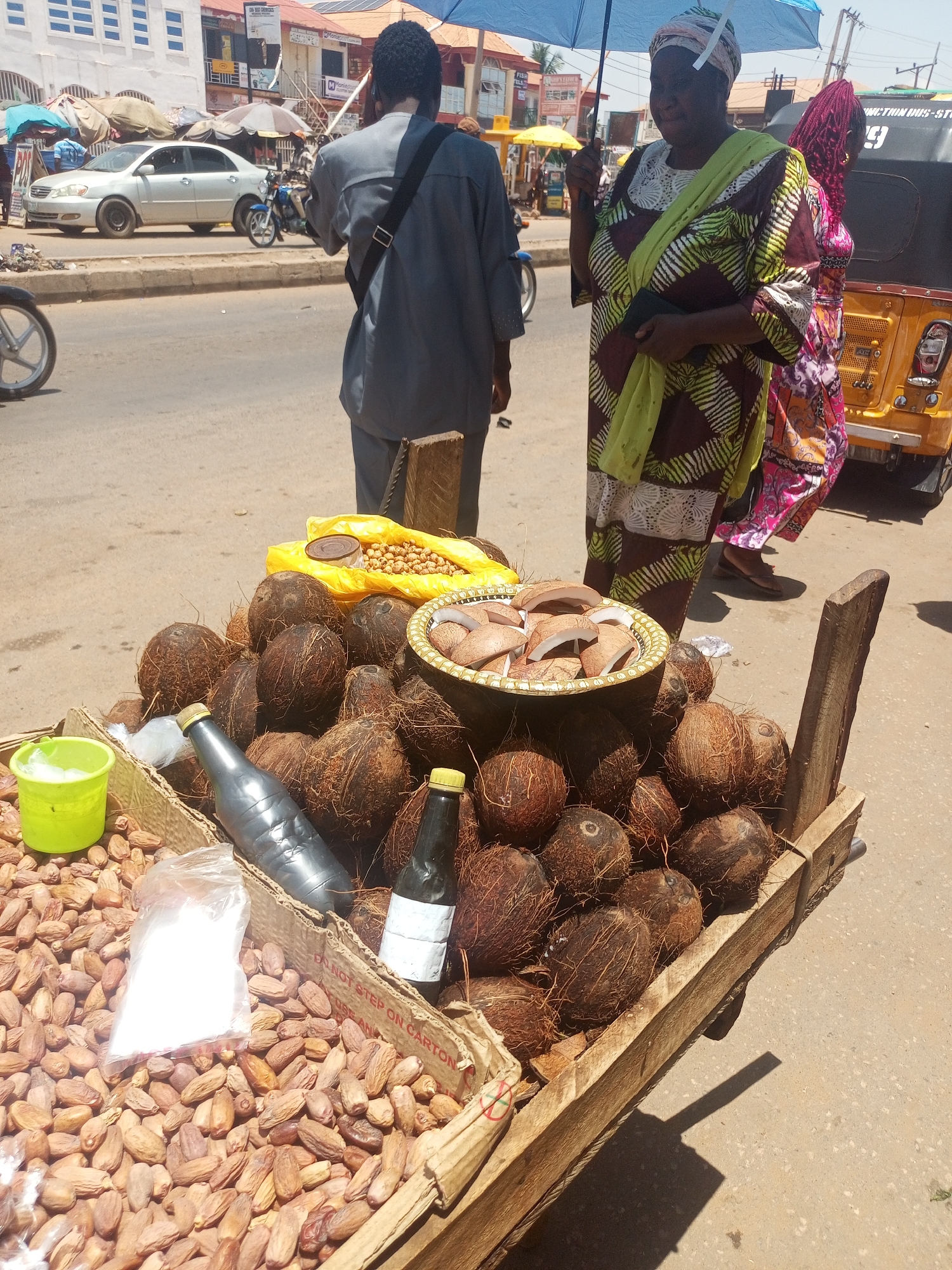 The image shows a street scene in Sabon Tasha, Nigeria, with a vendor's wooden cart filled with coconuts, dates, and a yellow bag of what appears to be chickpeas. A metal bowl filled with halved coconut pieces sits atop the pile of whole coconuts, along with two bottles of liquid on the cart. A small green bucket sits amidst the dates. A man in a gray shirt and black shoulder bag is walking away from the cart, seemingly on his phone, while a woman in a patterned dress holds an umbrella and walks next to the cart. Part of a yellow tricycle is visible to the right of the woman. In the background, there is a street with cars and buildings.