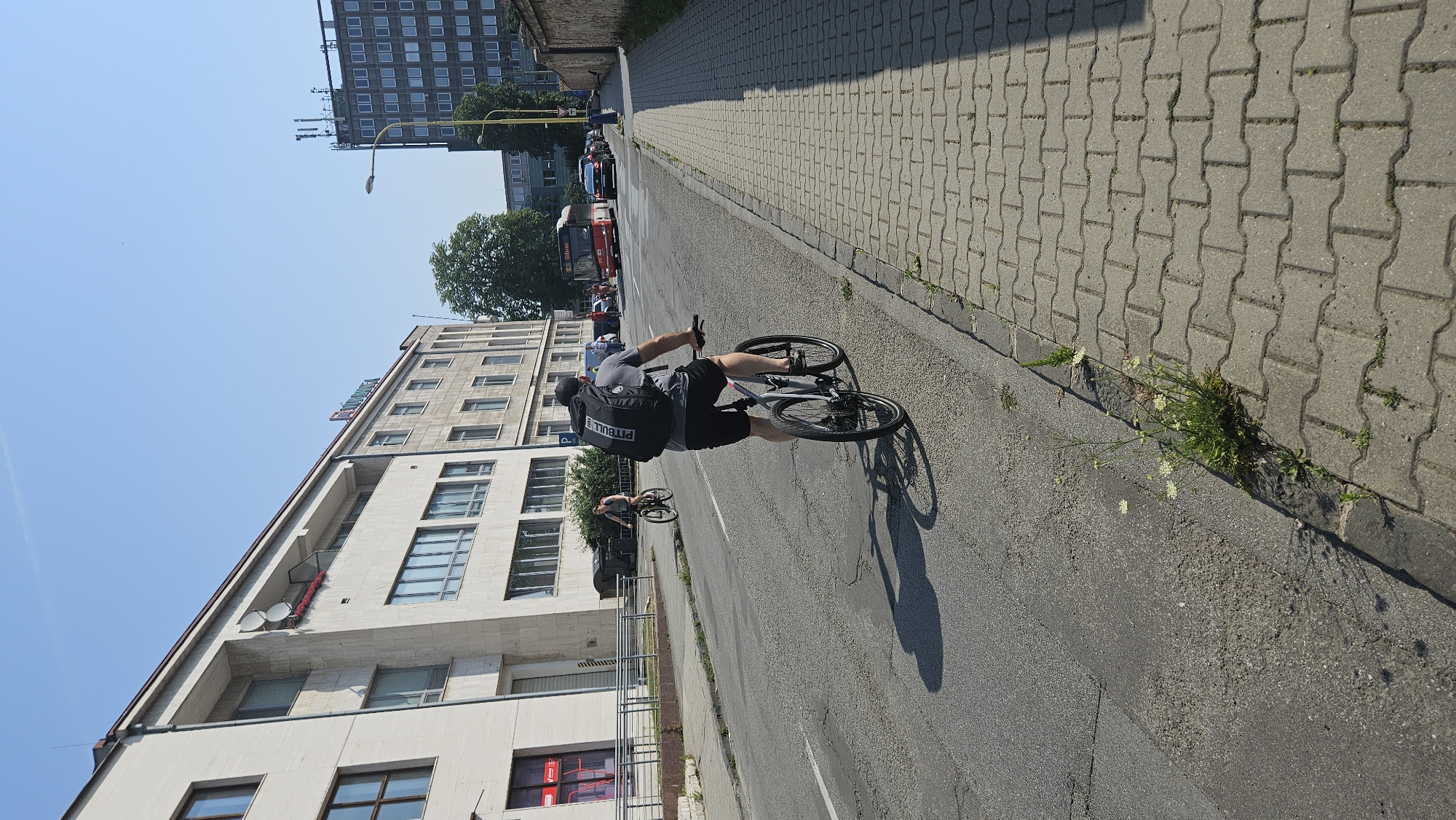 The image depicts a street scene in Prešov, Slovakia, under a clear blue sky. In the foreground, a man wearing a black baseball cap and a grey t-shirt is riding a bicycle. He has a large black backpack with "PITBULL" written on it. He is riding in the right lane of a two-lane road, with a sidewalk made of interlocking pavers to his right. The road surface is asphalt and shows signs of wear. To the left of the road is a sidewalk, and beyond that, a large, modern-looking building with a light-colored facade and numerous windows. Further down the street, another person is riding a bicycle. In the background, there are other buildings, including a taller, dark-colored building with many windows and a red bus is visible. Cars are also present on the road. The overall environment suggests a typical urban setting.