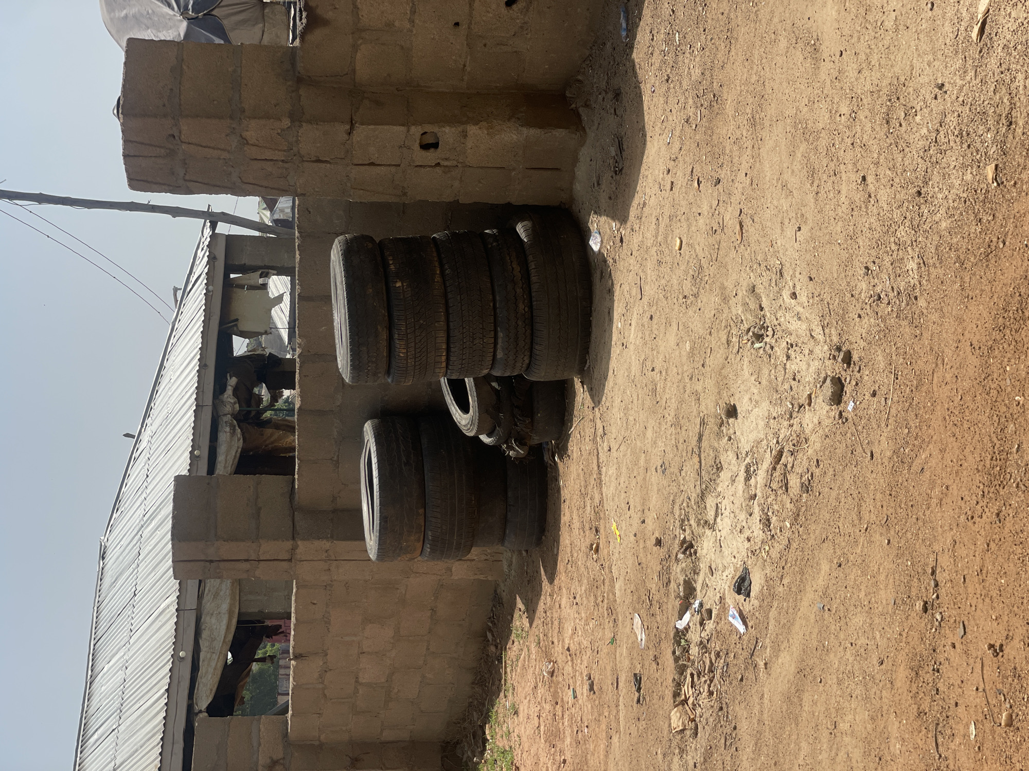 The image depicts an outdoor scene, likely a workshop or a storage area, set against a backdrop of a building constructed with cinder blocks. The primary subjects are a stack of several used tires, leaning against the wall. The tires appear worn, suggesting they are old or have seen significant use. To the left of the tires, part of a corrugated metal roof and a cinder block structure are visible, indicating some form of shelter or construction. A thin wooden pole with wires extending from it stands upright in the background. The ground is covered in dry, dusty earth, with scattered debris such as small pieces of paper and natural elements like twigs and small stones. The lighting suggests it is daytime, possibly late morning or early afternoon, with clear sky visible in the upper left corner. The overall impression of the scene is one of a functional, perhaps utilitarian, outdoor space in a location with arid or semi-arid conditions. There is no clear activity or event taking place beyond the static display of the tires. No people are clearly visible, and there are no discernible text elements in the image. The context provided suggests this location is Bauchi, Nigeria.