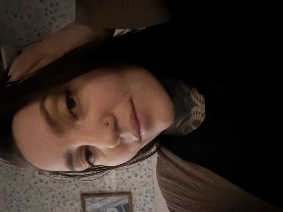 A selfie of a woman with dark hair and fair skin, positioned at an angle. Her head is slightly tilted. She has a nose piercing, and tattoos are visible on her neck. She is wearing a black shirt. The background includes a patterned wall and a partially visible framed picture. The lighting is dim, casting shadows on the left side of her face and on her shirt.