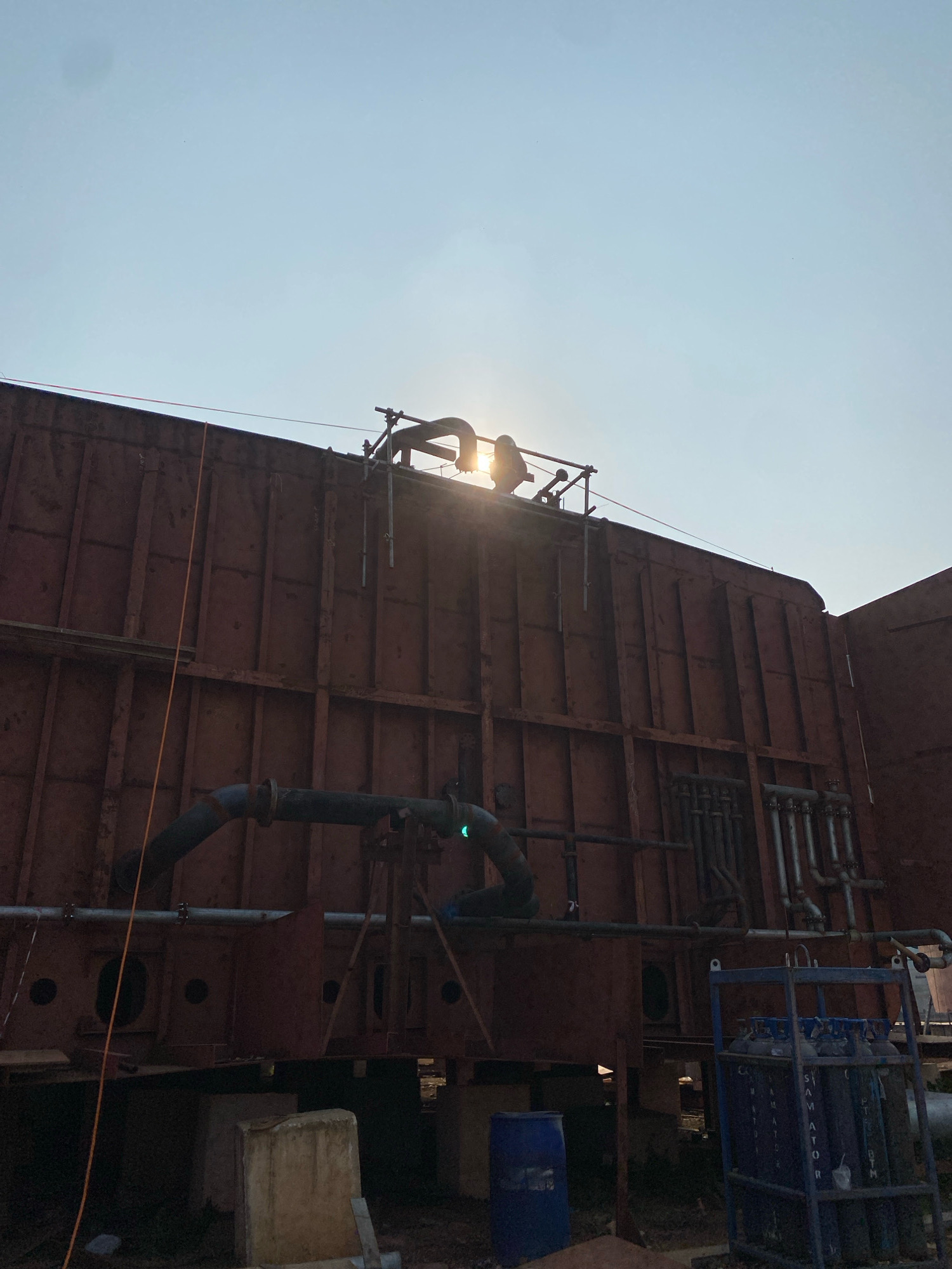 The image shows a large, rust-colored industrial structure, likely a section of a ship or a factory building, under construction or maintenance. The structure is supported by concrete blocks and is partially obscured by scaffolding. Several large pipes run along the exterior of the structure, some of which are dark gray and others appear to be made of stainless steel. A blue barrel and a metal cart containing several gas cylinders are visible at the base of the structure. The scene is set outdoors with a bright sky, and the sun is partially visible behind the structure, creating a lens flare effect. This scene is consistent with industrial areas within Batam, Indonesia, known for its shipbuilding and manufacturing sectors. There are no people clearly visible in the image.