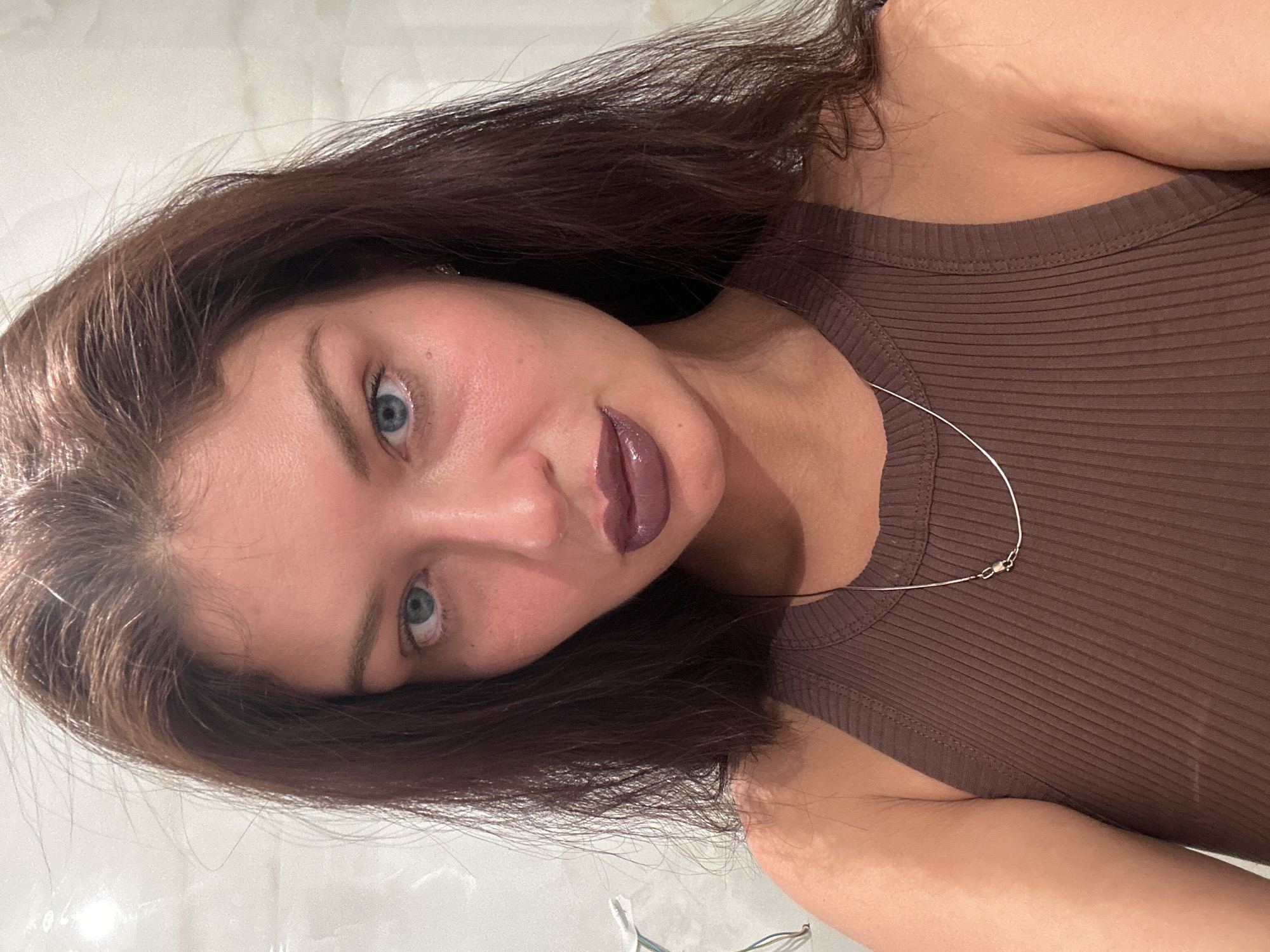 A woman with blue eyes and brown hair is seen in a close-up, selfie-style shot. She is wearing a ribbed, brown tank top and a thin, silver necklace. Her lips are painted a dark plum color. The background appears to be a blurred, bright white surface, possibly a wall. The location is not discernible from the image; there is no indication of Moscow, Russia.