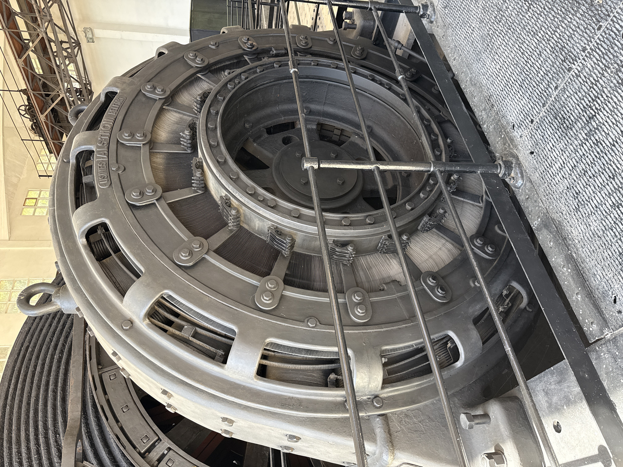 The image shows a large, industrial generator, likely an alternator, in a dimly lit hall with a concrete floor covered in a grid pattern. The generator is predominantly dark grey and metallic, with visible bolts, wires, and a large central rotor. The words "USINES ALSTHOM BELFORT" are inscribed on its casing. A metal railing surrounds the generator, separating it from the viewer. In the background, structural beams and machinery are visible, along with windows in a cream-colored wall. A large, dark, ribbed cylinder is partially visible to the left of the generator. This generator is located within the former hydroelectric power station of the Cardona Salt Mines in Cardona, Spain.
