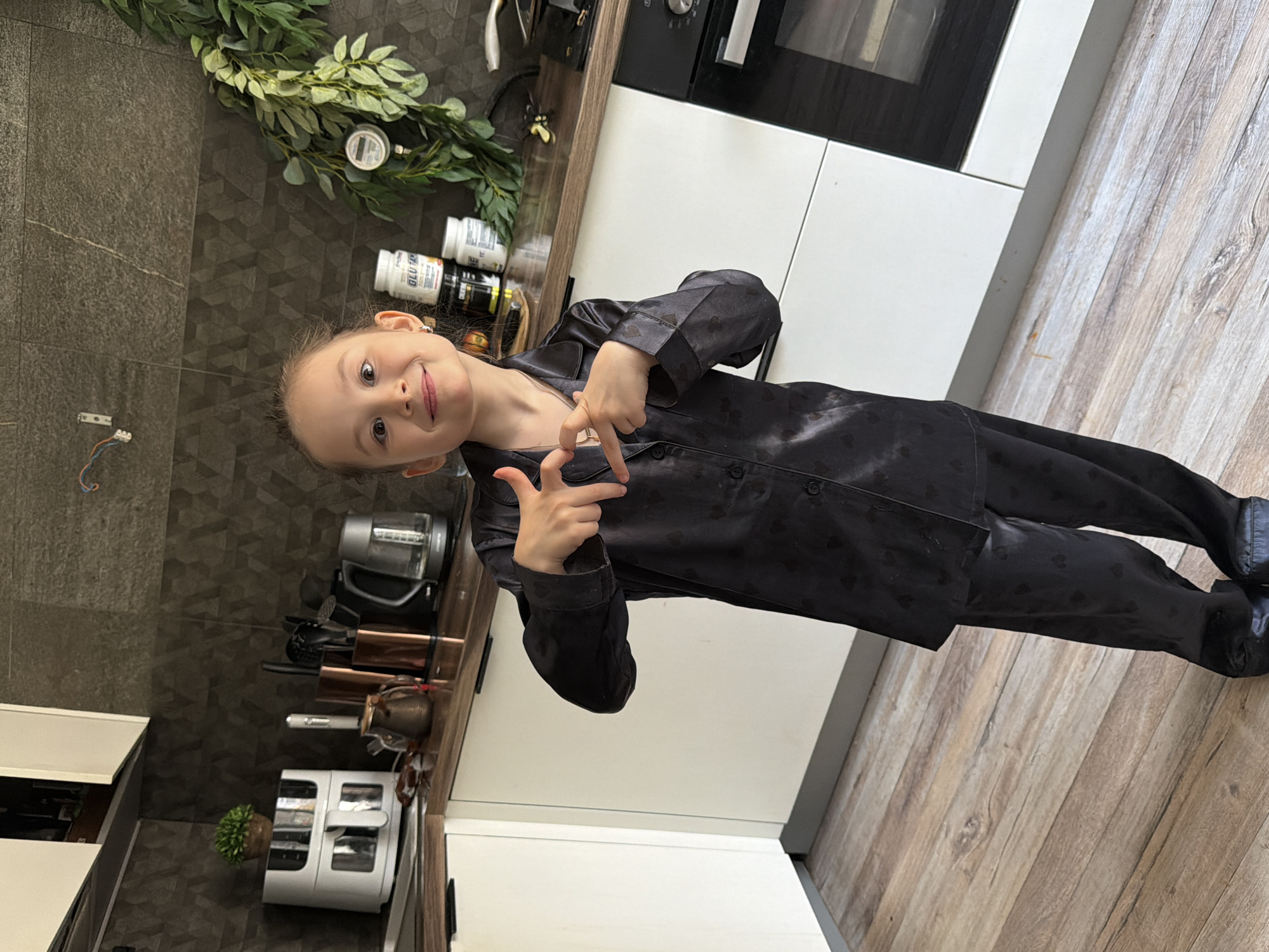 A young girl in black pajamas stands in a kitchen with wooden floors and dark tile walls. She is facing the camera and gesturing with both hands. Behind her is a countertop with various kitchen appliances and containers, including a toaster oven and a blender. A sprig of greenery sits on the countertop. The kitchen cabinets are white and black. The location appears to be a residential kitchen, but there is no specific evidence to confirm its location within the city of Perm, Russia.