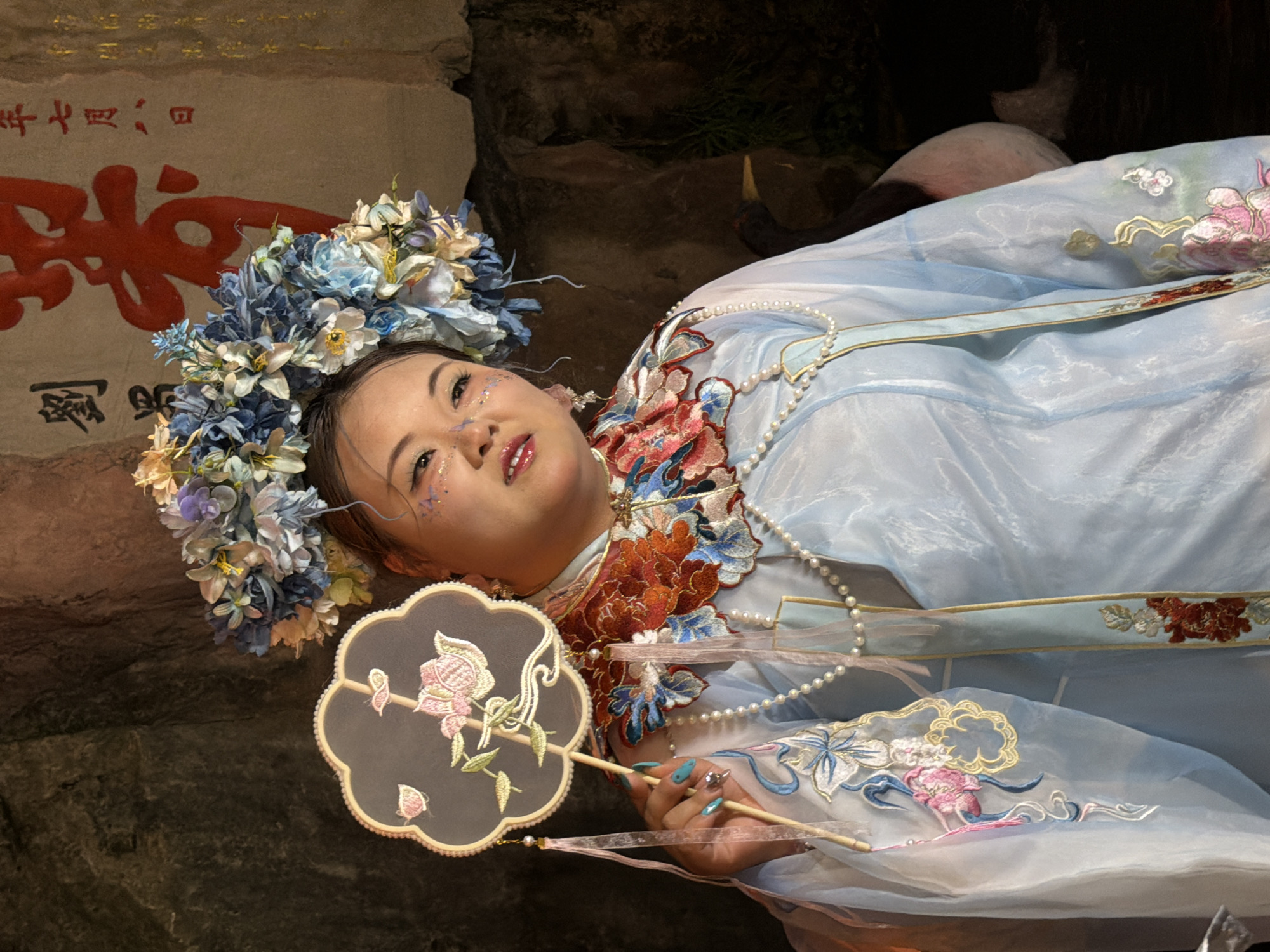 A person is posing for a photograph while holding a fan. The person is wearing a traditional Chinese outfit with intricate embroidery and a floral headdress. The background appears to be a natural setting with a rock formation and a painting with Chinese calligraphy. There is a bird partially visible in the background on the right side of the image. The lighting suggests an indoor setting, possibly a studio or a room with dim lighting. The scene appears to be in Yuzhong District, China, based on the presence of traditional Chinese elements.