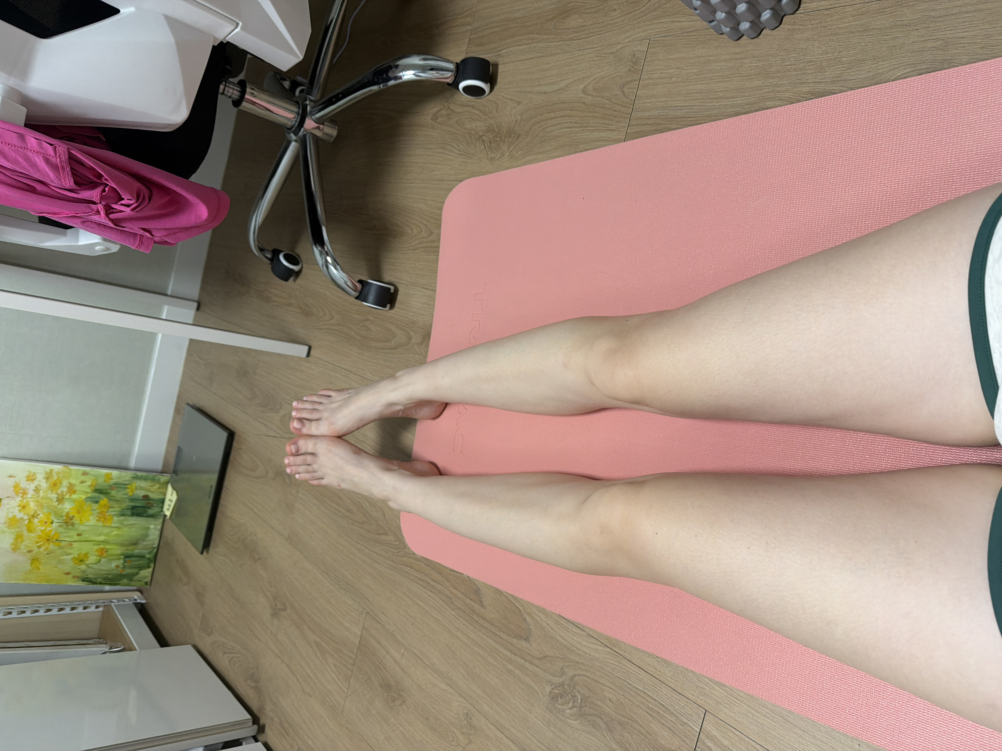 This media file is an overhead shot of a person's legs resting on a pink yoga mat. The scene is indoors, likely a home or studio, with a wooden floor visible. In the background, a white office chair with chrome accents and black wheels is partially visible. A pink garment, possibly athletic wear, is draped over the chair. To the left, a laptop and a painting of yellow flowers on a canvas are placed on the floor. A white shelf unit is also visible. The lighting suggests daytime. The person's legs are relaxed, with bare feet positioned at the top of the yoga mat, suggesting they are about to exercise or have just finished. There is no discernible text in the image.