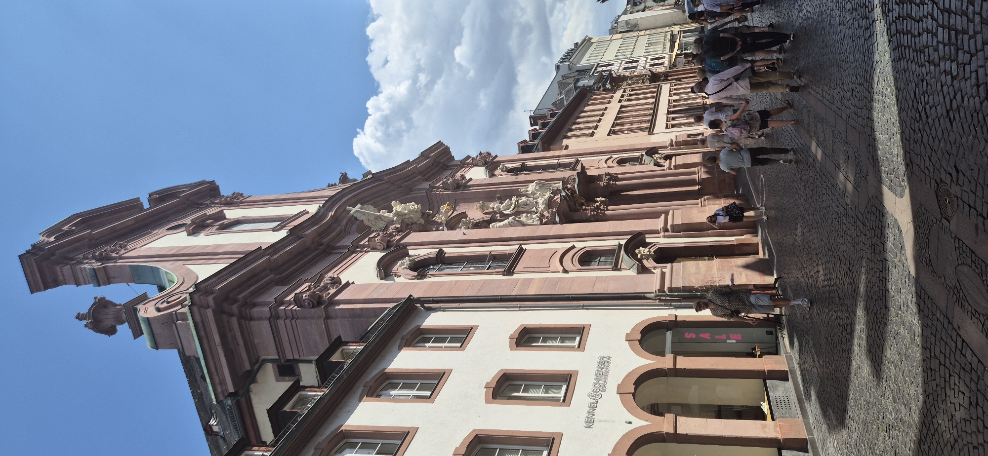 The image shows a street scene in Mainz, Germany, featuring a large, ornate building with pink sandstone facade and white accents, likely a church or historic public building, on the right. The building exhibits intricate architectural details, including sculptures, columns, and decorative cornices. To the left of this building is a lighter-colored structure with arched entrances on the ground floor, one of which has a "SALE" sign visible. The street is paved with cobblestones and is populated by several individuals walking in various directions, suggesting it is a public thoroughfare. The sky is blue with scattered clouds, and sunlight casts shadows on the cobblestone pavement, indicating it is daytime. The overall atmosphere is that of a bustling European city street with historical architecture.