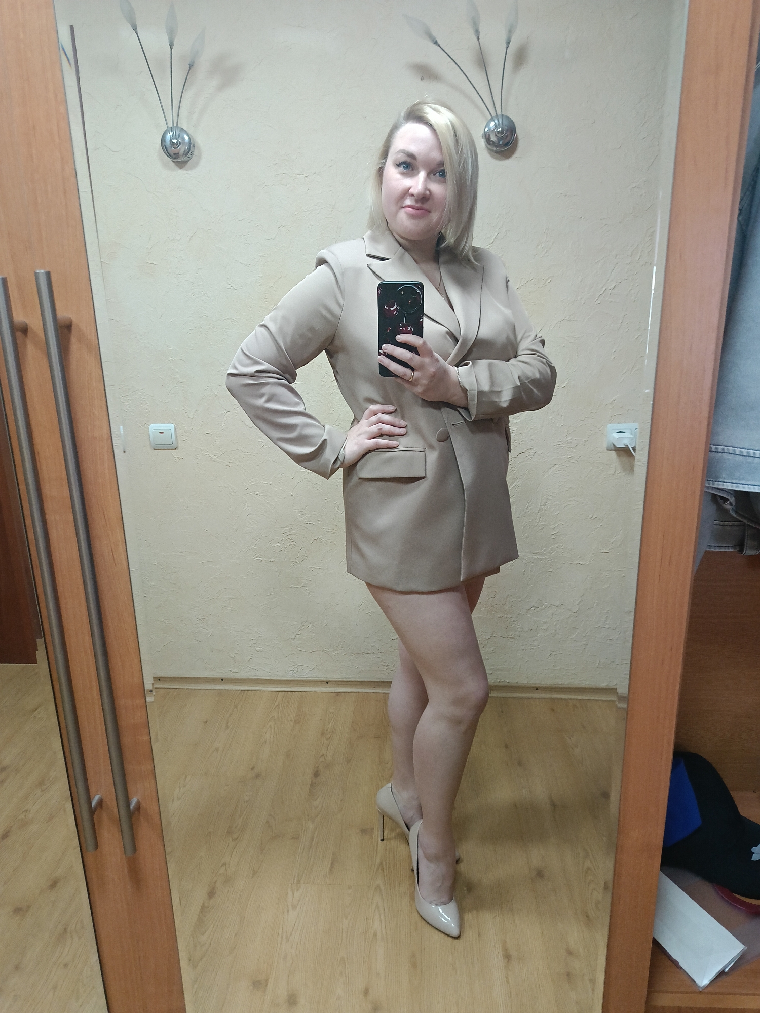 A full-length, indoor photograph shows a woman standing in front of a mirror, taking a selfie. She is wearing a beige blazer dress and beige high-heeled shoes. She has short, blonde hair. She is holding a phone with a black case. The room has beige walls, a light-colored wooden floor, a light switch, and a wall sconce. Part of a wooden wardrobe is visible on either side of the mirror. The location is Chaikovsky, Russia.