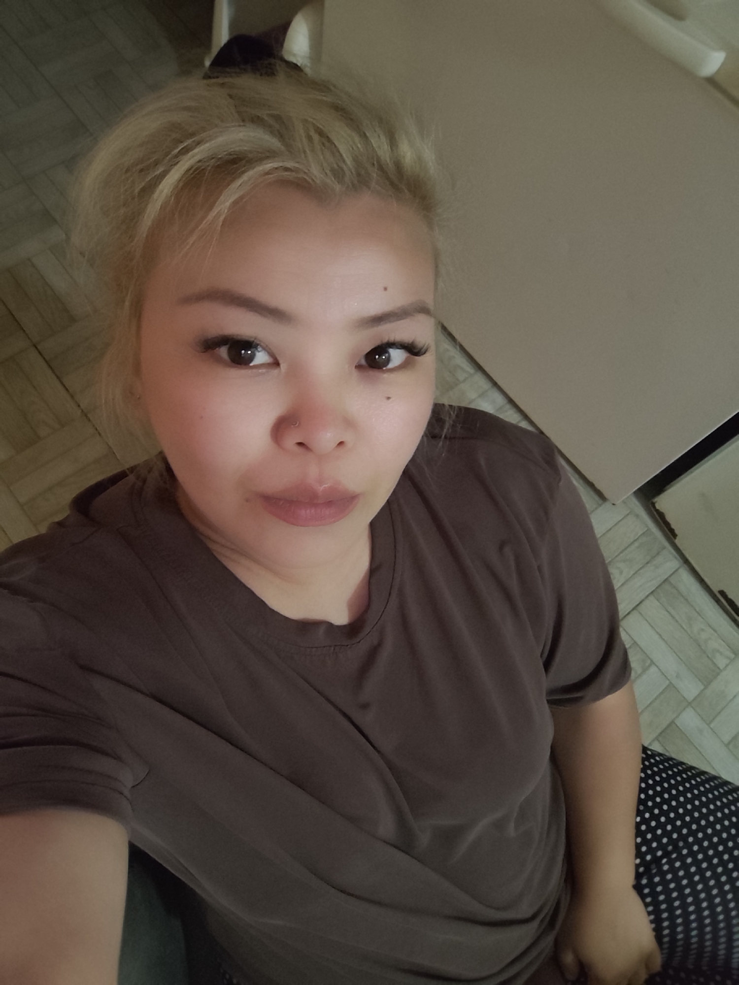 An adult female is captured in a close-up, high-angle selfie. She has light-colored, possibly bleached or dyed, blond hair styled loosely, with some strands falling over her forehead. Her dark eyes gaze directly at the camera, accentuated by prominent eyelashes. She has a small stud piercing on her left nostril. Her skin appears smooth, and her lips are slightly parted in a neutral expression, with a subtle natural lip color.
She is wearing a plain, dark brown, short-sleeved t-shirt. Below her waist, the pattern of dark pants with white polka dots is visible. Her left arm is slightly raised, suggesting she is holding the camera for the selfie.
The background is a simple indoor setting. To her right, a light-colored, possibly off-white or beige, wall or large surface occupies the upper part of the frame. The floor beneath her features light-colored, rectangular planks arranged in a geometric pattern, possibly a parquet or laminate style. The lighting is soft and even, typical of indoor artificial light, with no harsh shadows indicating direct sunlight. No text is visible in the image. The overall impression is a casual, posed portrait. While the location context of Sarıyer, Turkey is provided, no specific visual cues within the image confirm this.