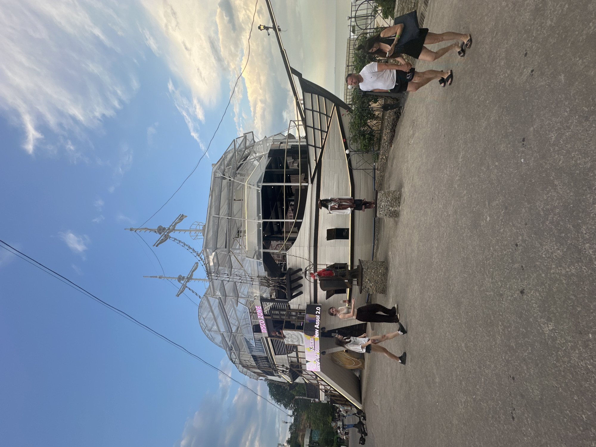 A group of people are gathered near a large, white, boat-shaped structure with multiple levels and a partially open-air design. The structure resembles a pirate ship, with masts, rigging, and what appears to be a "DISCO ZONE" sign. There are neon lights on the building, one of which reads "New Assol 2.0". Some people are walking on a paved area next to the structure, while others are standing or interacting on the lower levels of the boat-like building. The scene appears to be outdoors, with a blue sky and scattered clouds visible, suggesting it could be daytime or early evening. The overall atmosphere seems casual and social.