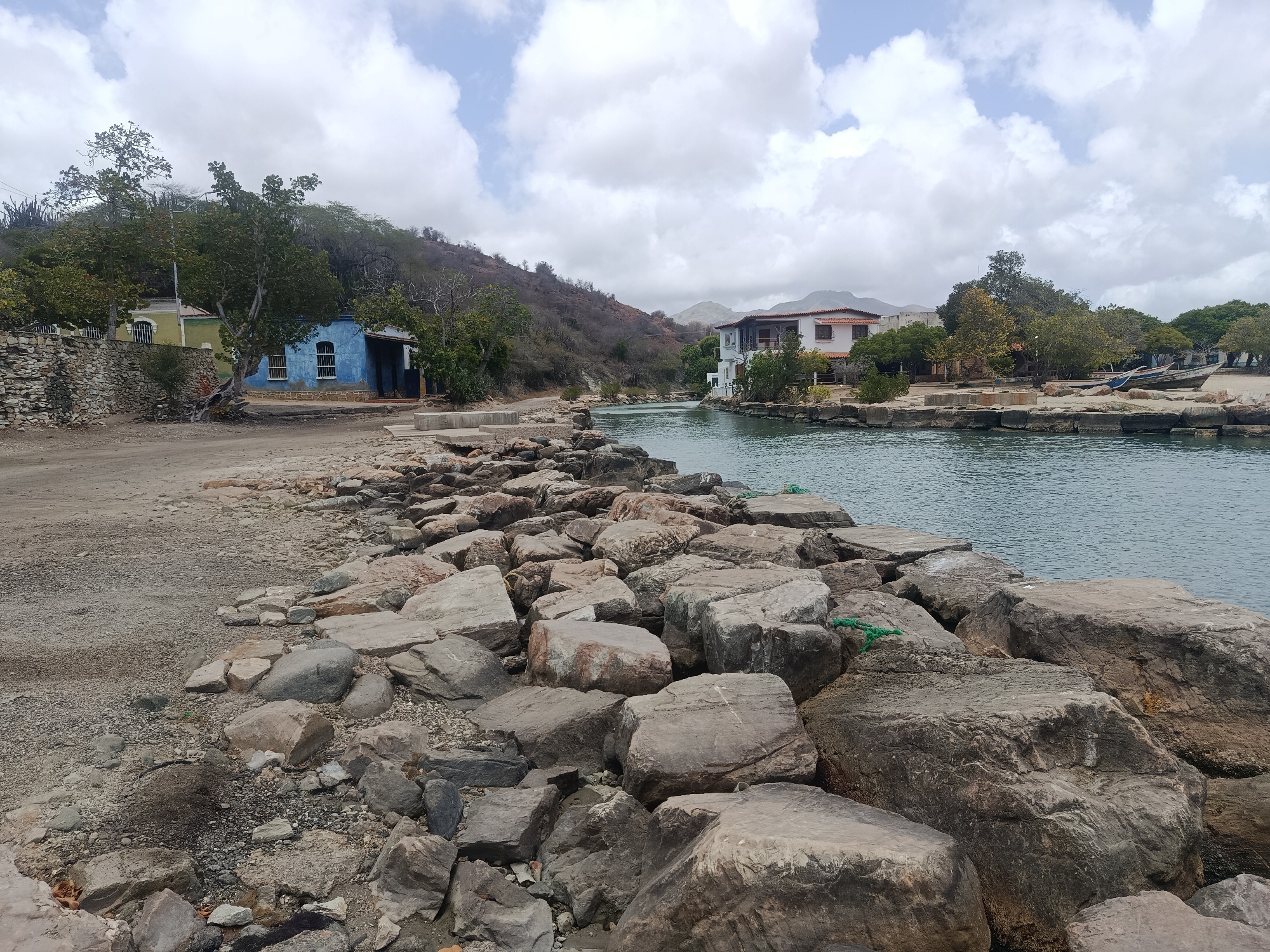 The image shows a waterfront view in Pedro González, Venezuela, with a rocky breakwater in the foreground leading to a channel. A mix of light-colored and gray rocks make up the breakwater. The calm water channel extends between shorelines, and is flanked by sparse vegetation. Two buildings are visible on the shorelines: one painted blue on the left, and a larger white building with an orange roof on the right. A boat is also seen near the shore to the right of the frame. Beyond the buildings and the channel, a small hill with little foliage is visible and in the distant background are mountainous peaks under a cloudy sky. The weather appears overcast.
