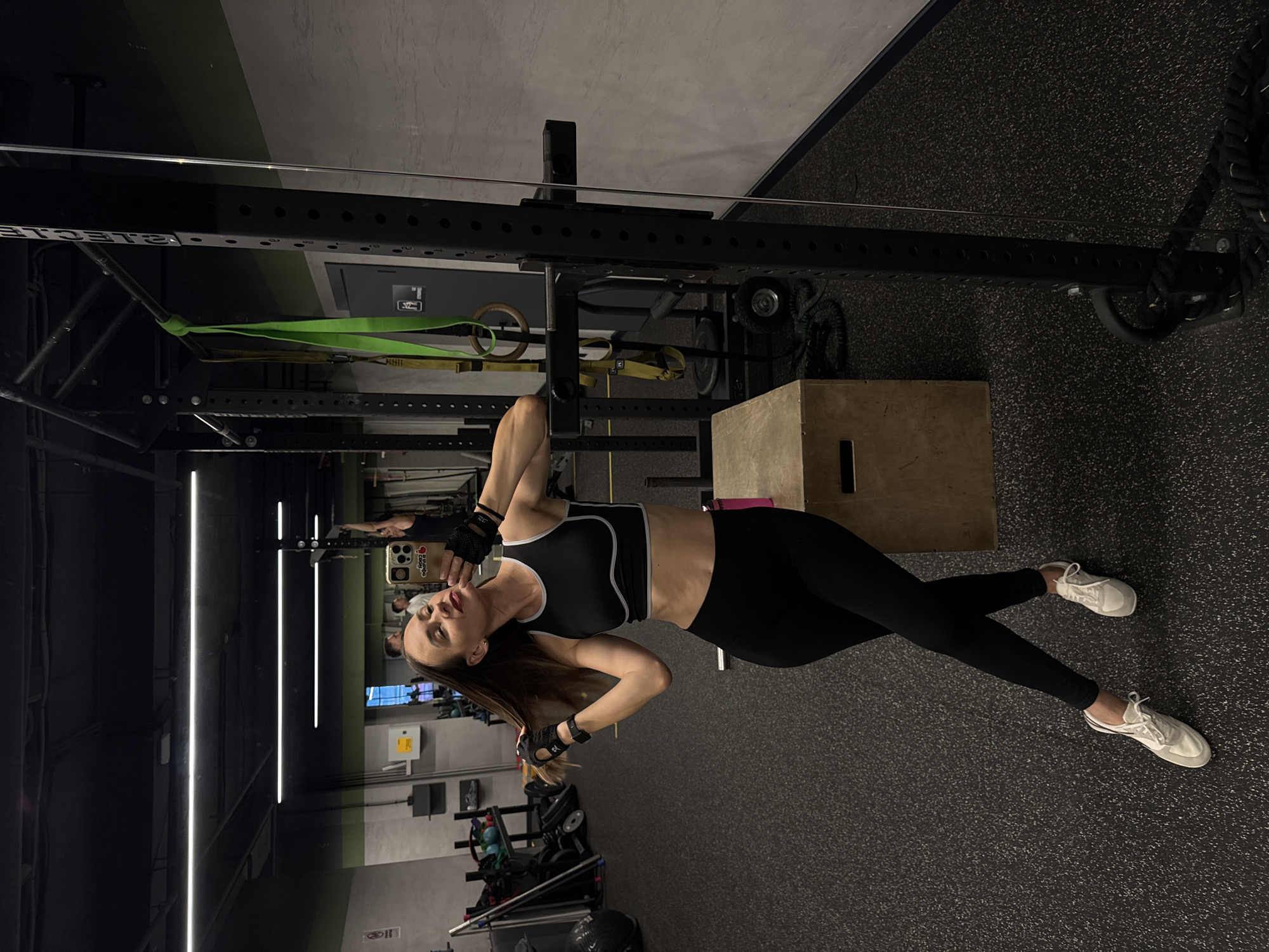 A woman stands in a gym and takes a selfie in a mirror. She is wearing a black sports bra and black leggings, white athletic shoes, and black fingerless gloves. Her hair is long and brown, and she has red lipstick on. She is leaning on a black weight rack and holding her hair with one hand while posing with the other. A pink water bottle is on a wooden box behind her, and other gym equipment is visible in the background. The gym appears to be a modern facility with black rubber flooring and bright overhead lighting. There are other people visible in the reflection in the mirror, as well as gym equipment like weight racks, ropes, and a plyometric box. The overall impression is one of a woman preparing for or finishing a workout in a well-equipped gym.