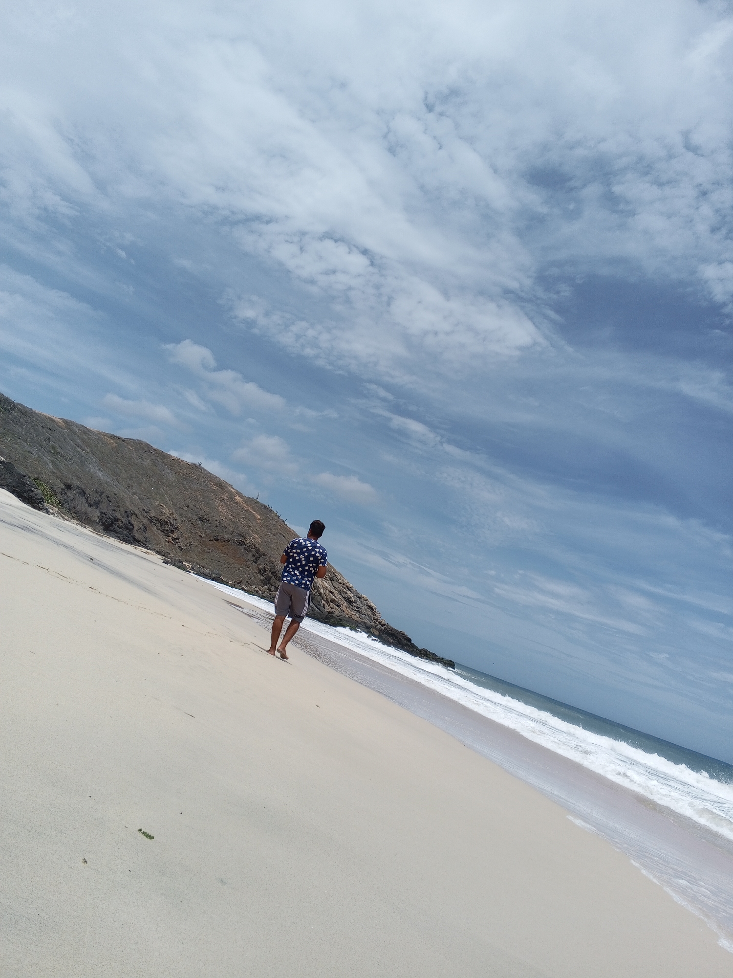 A man walks along a sandy beach towards the ocean. He is wearing a blue patterned shirt and grey shorts. The beach stretches along the edge of a hill or small mountain. The sky is mostly blue with streaks of white clouds. The location is a beach in Altagracia, Venezuela.