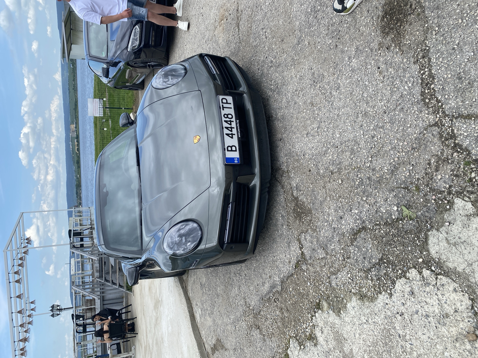The image shows the front of a gray Porsche parked on a concrete surface near a waterfront area. The license plate reads "B 4448 TP" and indicates the car is registered in Bulgaria. A black car is partially visible behind it, and two people are standing near the black car. A structure with hanging lights is to the left, with two people sitting below it. The body of water appears to be a lake or wide river. The location is likely Konstantinovo, Bulgaria based on contextual data. The weather is sunny with a partly cloudy sky. The scene appears casual, possibly a gathering near a lakeside restaurant.