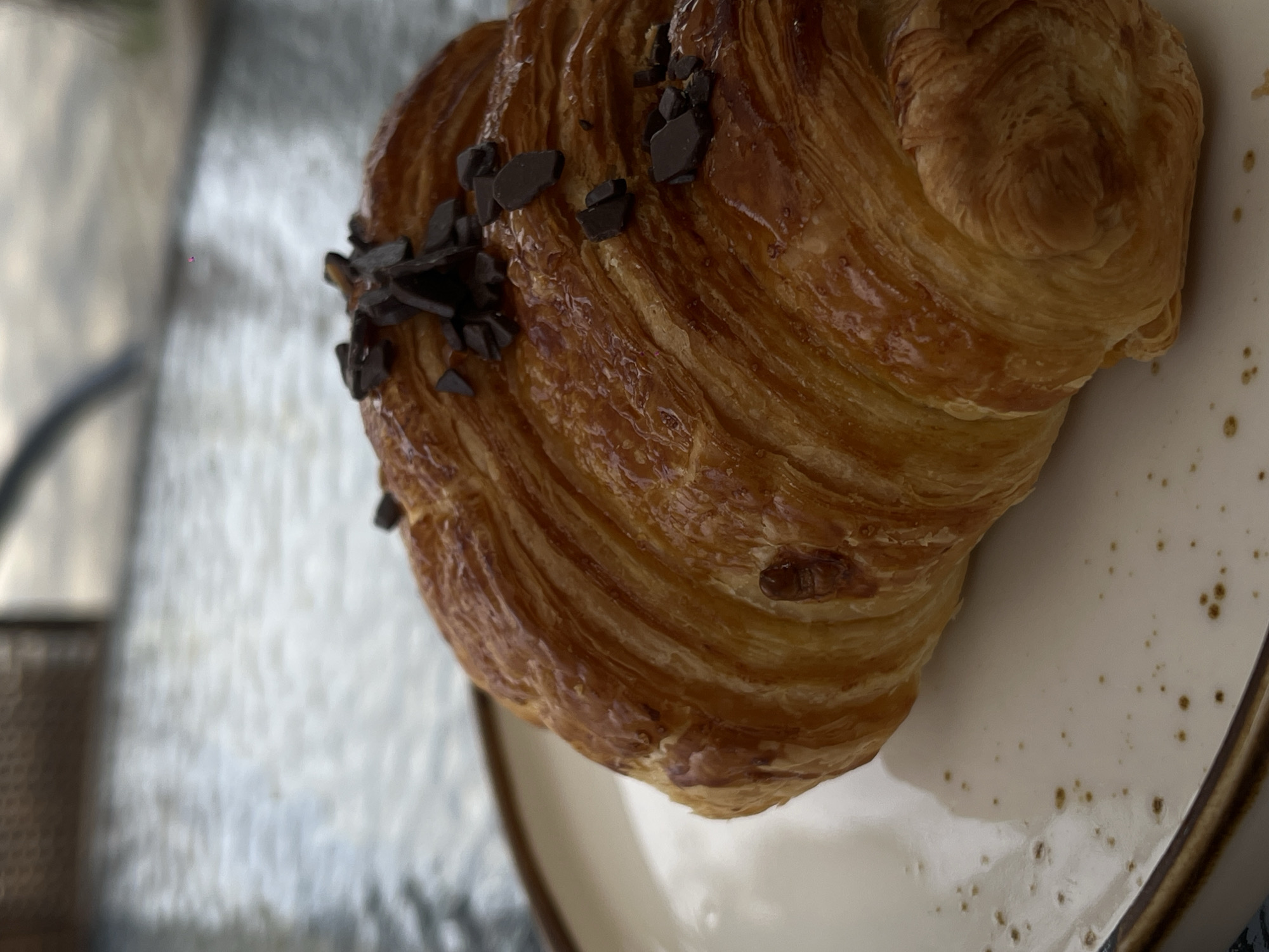 The image shows a chocolate croissant on a white plate with brown speckles. The croissant is golden brown and flaky, with chocolate shavings on top. The background is blurred, but appears to be a table. The location is not identifiable from the image.