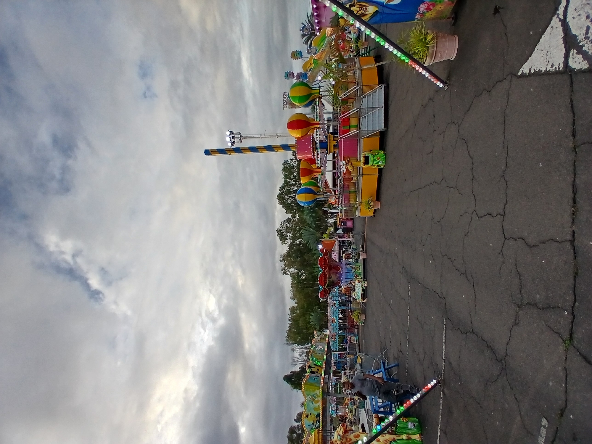 The image displays an outdoor amusement park or carnival operating under an overcast sky in Ciudad Guayana, Venezuela. The ground is dark gray asphalt pavement, extensively cracked, with faint white line markings.
In the foreground, a diagonal structure with a strip of colored LED lights (red, green, white) extends from the lower left. To the left of the center, a male individual in a grey t-shirt and dark pants stands, looking down at a mobile phone. He is positioned near several blue plastic chairs and a low-lying carnival ride featuring a yellow animal head, possibly a giraffe.
Multiple carnival rides occupy the mid-ground and background. A prominent ride on the right features a tall, yellow and blue striped central tower with floodlights at its peak. Surrounding its base are four hot-air balloon-themed gondolas (one blue/yellow, one red/yellow, two green/yellow). This ride has a yellow platform, a metal stair access, and a green animal-shaped trash bin. A sign above this ride reads "TABOGA." Further to the left, another large, multi-colored spinning ride with animal figures is visible. Other assorted rides and structures are scattered throughout the background.
Metal barriers define ride perimeters, and potted plants serve as decor near some attractions. Trees and foliage line the distant background beneath a sky dominated by grey clouds, with some brighter areas indicating diffused light penetration. Several rides exhibit illuminated decorative lights and operational floodlights.