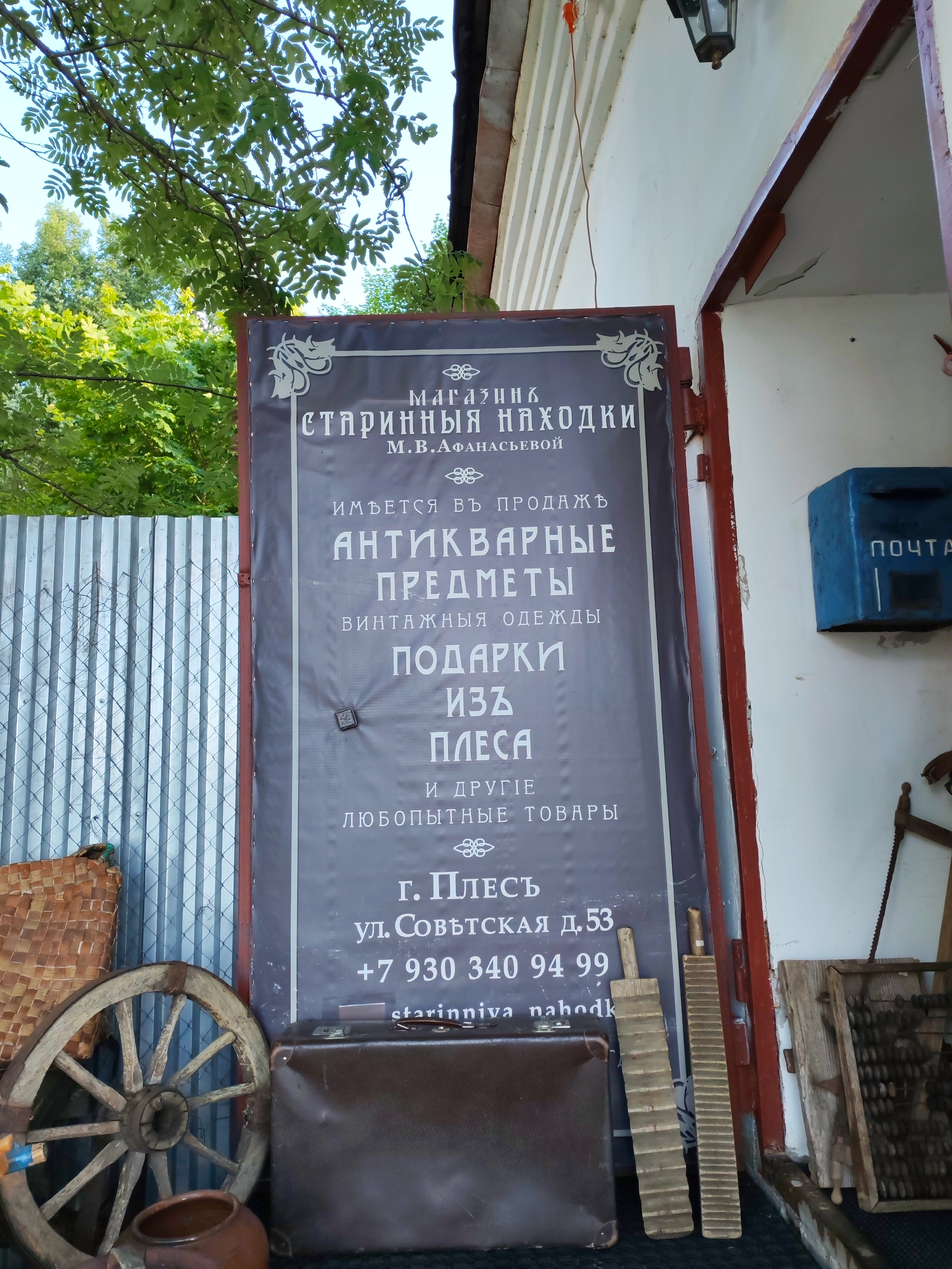 This is an outdoor, eye-level, medium shot of a storefront in Plyos, Russia. The focus is on a large, dark sign advertising an antique shop. The sign is ornate, with decorative borders and Cyrillic text in white. It reads "МАГАЗИН СТАРИННЫЯ НАХОДКИ М.В. АФАНАСЬЕВОЙ" which translates to "Shop of Old Finds M.V. Afanasyeva". Below this, it lists items for sale: "АНТИКВАРНЫЕ ПРЕДМЕТЫ" (Antique items), "ВИНТАЖНЫЯ ОДЕЖДЫ" (Vintage clothing), "ПОДАРКИ ИЗЪ ПЛЕСА" (Gifts from Plyos), and "И ДРУГІΕ ЛЮБОПЫТНЫЕ ТОВАРЫ" (And other curious goods). The address is given as "Г. ПЛЕСЪ УЛ. СОВѢТСКАЯ Д. 53" (Plyos, Sovetskaya St. 53), followed by a phone number: "+7 930 340 94 99". A website, "starinniva nahodk," is also partially visible.
To the left of the sign, a portion of a corrugated metal fence is visible, along with a woven basket and a rustic wooden wagon wheel. A dark brown suitcase and a clay pot sit in front of the sign. To the right of the sign, a blue mailbox with the word "ПОЧТА" (Post) is mounted on a white wall. Further to the right, part of a wooden structure, an old abacus, and a saw are visible. Above the sign, a black outdoor lantern hangs from an overhang. The scene appears to be set during the daytime, with dappled sunlight filtering through the green leaves of trees in the upper left of the frame. The overall impression is one of a quaint, old-fashioned shop selling vintage and antique items.