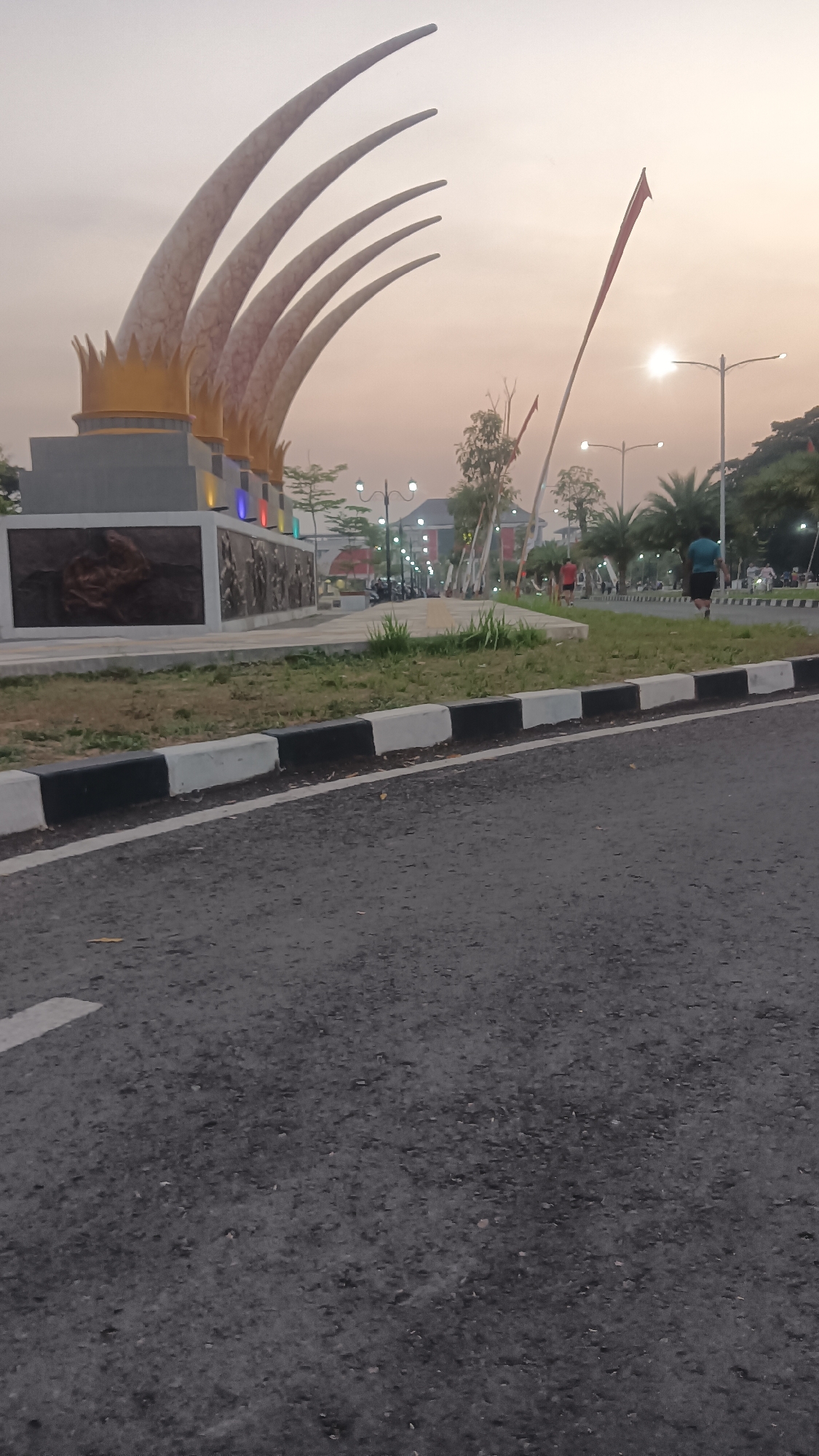 The image depicts a public area at dusk, likely in Sragen Kulon, Indonesia. A prominent monument with tall, curved elements resembling golden crowns and sharp, pointed tips dominates the left side of the frame. The monument is situated on a concrete base adorned with relief carvings and colorful lights.
In the background, a street is visible with several lampposts illuminated, suggesting an evening or twilight setting. A flag pole with a red flag is also present. A paved road with a distinct black and white painted curb runs in the foreground, taking up the lower half of the image.
Several individuals are present in the scene. Two people are jogging or running on a path alongside the monument and road. Other figures can be seen further in the background, appearing to be walking or standing, engaged in various activities. Trees and some vegetation are scattered throughout the area, contributing to the park-like atmosphere. The sky is a soft, pale orange, indicative of sunset or sunrise.