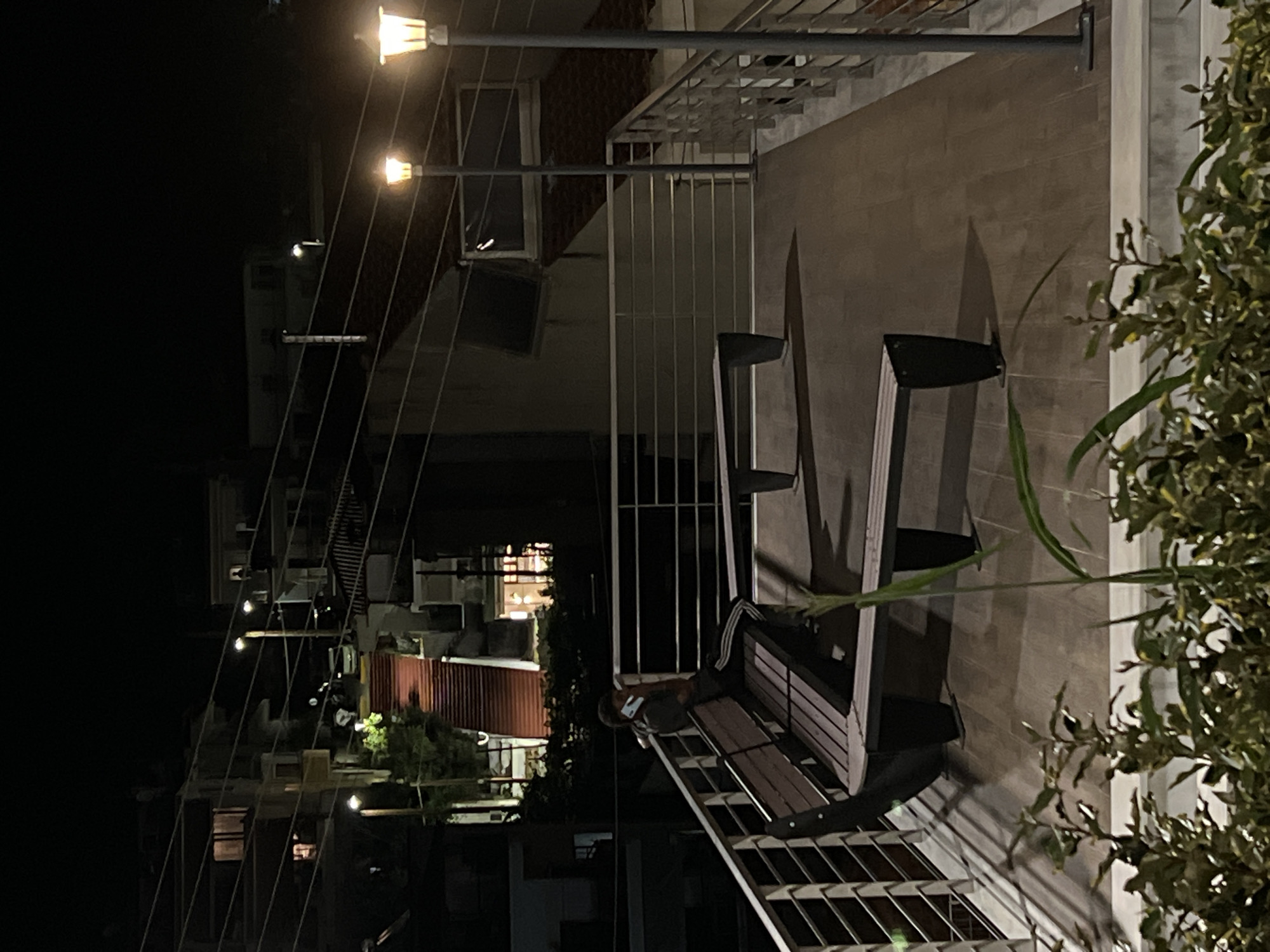 At night, a person is sitting on a wooden bench on a rooftop patio in Sithonia Municipal Unit, Greece. The person is wearing dark clothing with white stripes on the pants and is holding a mobile phone to their ear, engaged in a conversation. Two street lamps with warm light illuminate the patio. In the background, buildings are visible with some lights on in the windows. A red staircase ascends a nearby hillside. Lush green foliage frames the bottom of the image.
