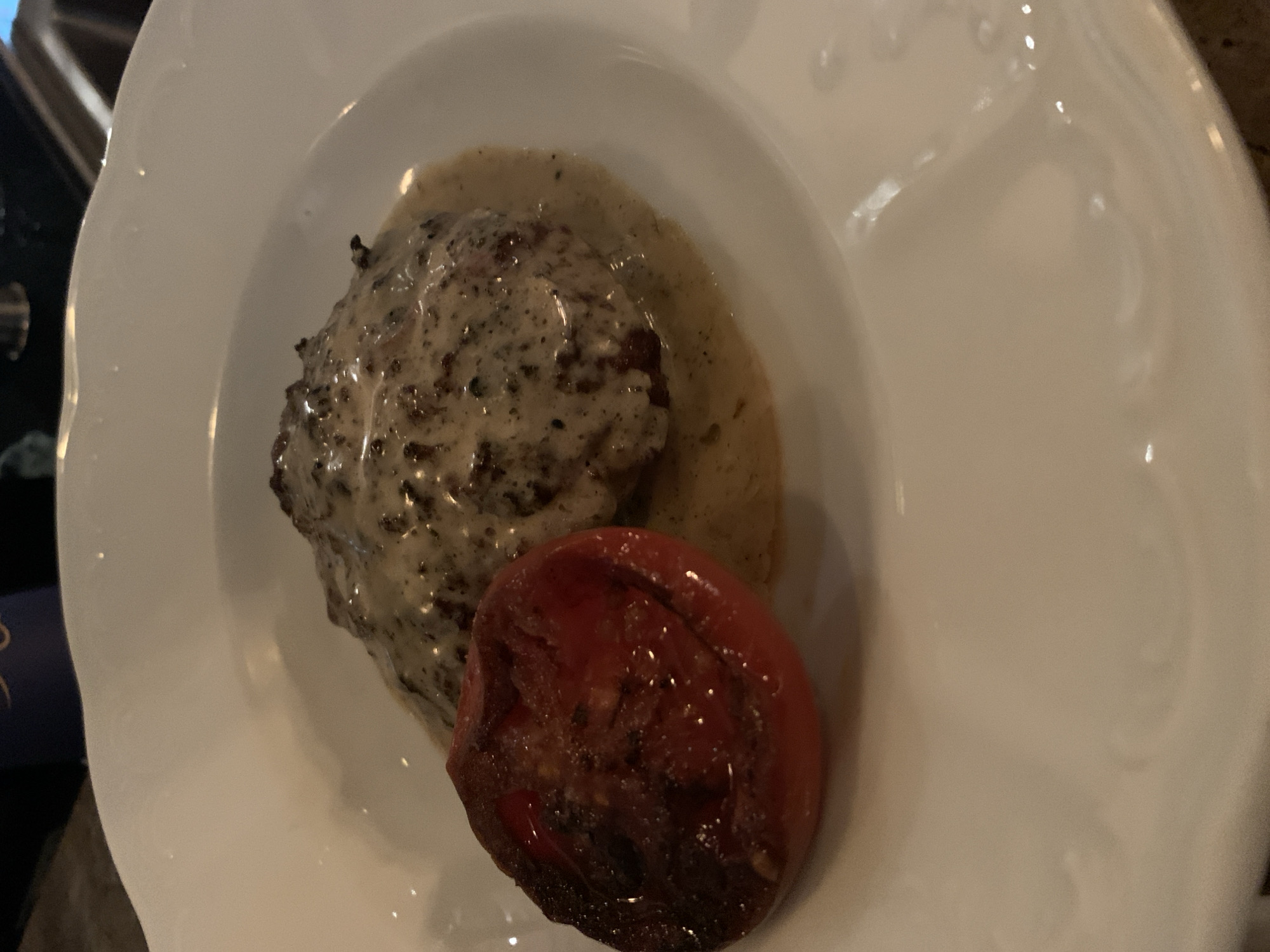 The image presents a close-up view of a single serving of food on a white, decorative plate, captured from an overhead perspective. The dish features a main component, likely a meat patty or small steak, generously covered in a rich, creamy, light brown sauce. This sauce is speckled with numerous dark particles, suggesting ingredients like ground pepper or finely chopped mushrooms.
Positioned alongside the sauced meat is a single, roasted or grilled tomato half. It exhibits a deep red hue with some darker, slightly charred areas and a glossy, softened appearance. The white plate has an ornate, embossed pattern along its rim, adding a touch of elegance to the presentation.
The background is dark and out of focus, indicating an indoor setting, likely a restaurant or a dinner table. The lighting is dim and warm, creating a cozy and inviting ambiance around the meal. No individuals are visible in the frame, and there is no discernible text. The image captures a meal ready to be consumed. The location context provided is Tel-Aviv, Israel.