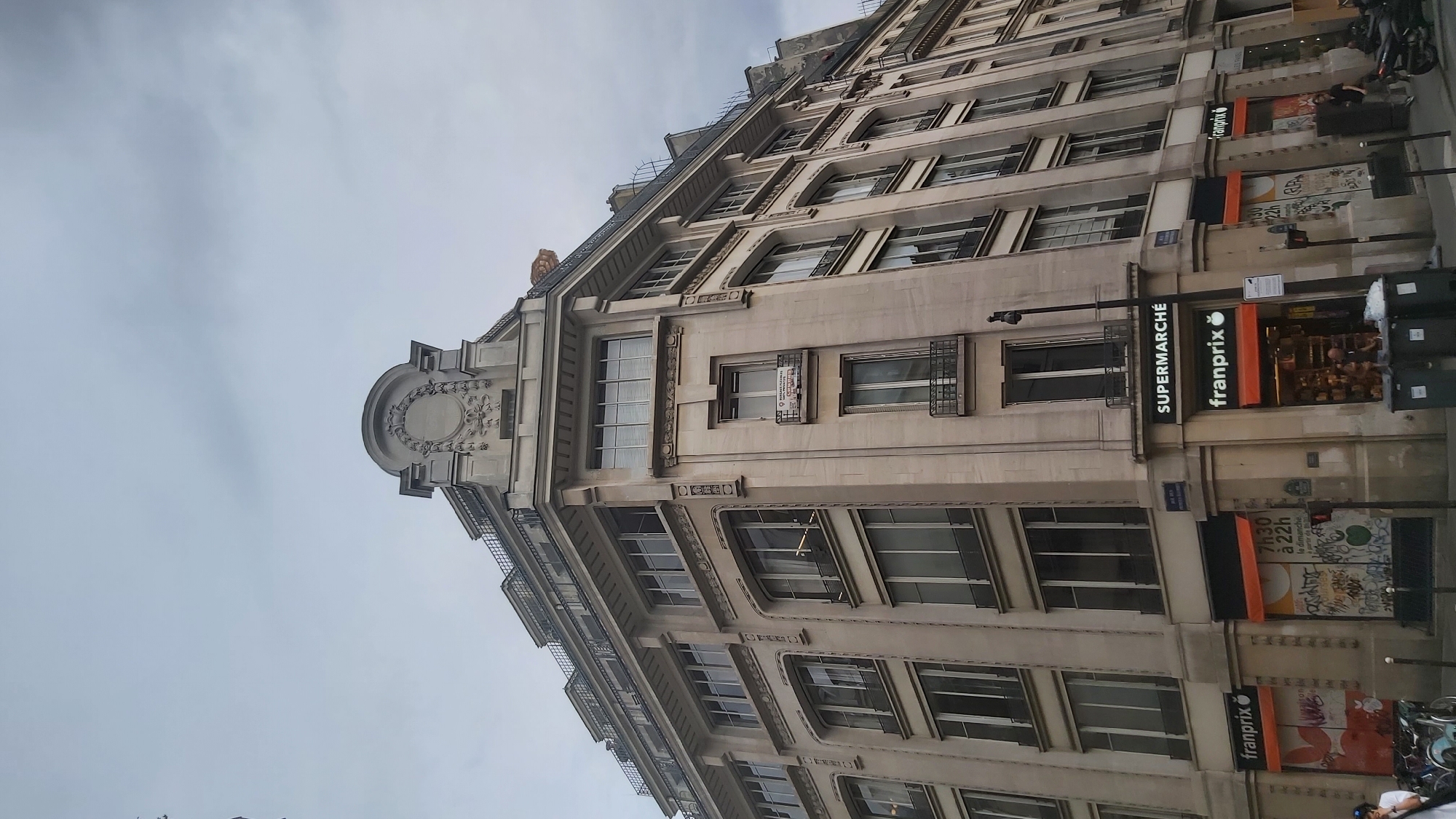 The image shows a street-level view of a multi-story building in Paris, France, on an overcast day. The building is light-colored stone with numerous windows, some with blinds, and decorative architectural elements. The ground floor features two Franprix convenience stores, one on the corner and another adjacent to it. The corner store has an open entrance displaying merchandise and a person visible inside. The adjacent store has its windows covered with graffiti and an orange awning. A street sign above the stores indicates "Rue de ...". To the left, there are bicycles parked, and a person in a white shirt is visible. To the right, a motorcycle is parked, and another person is walking away. Trash bins are placed on the sidewalk in front of the stores. The sky is cloudy, creating a diffused light.