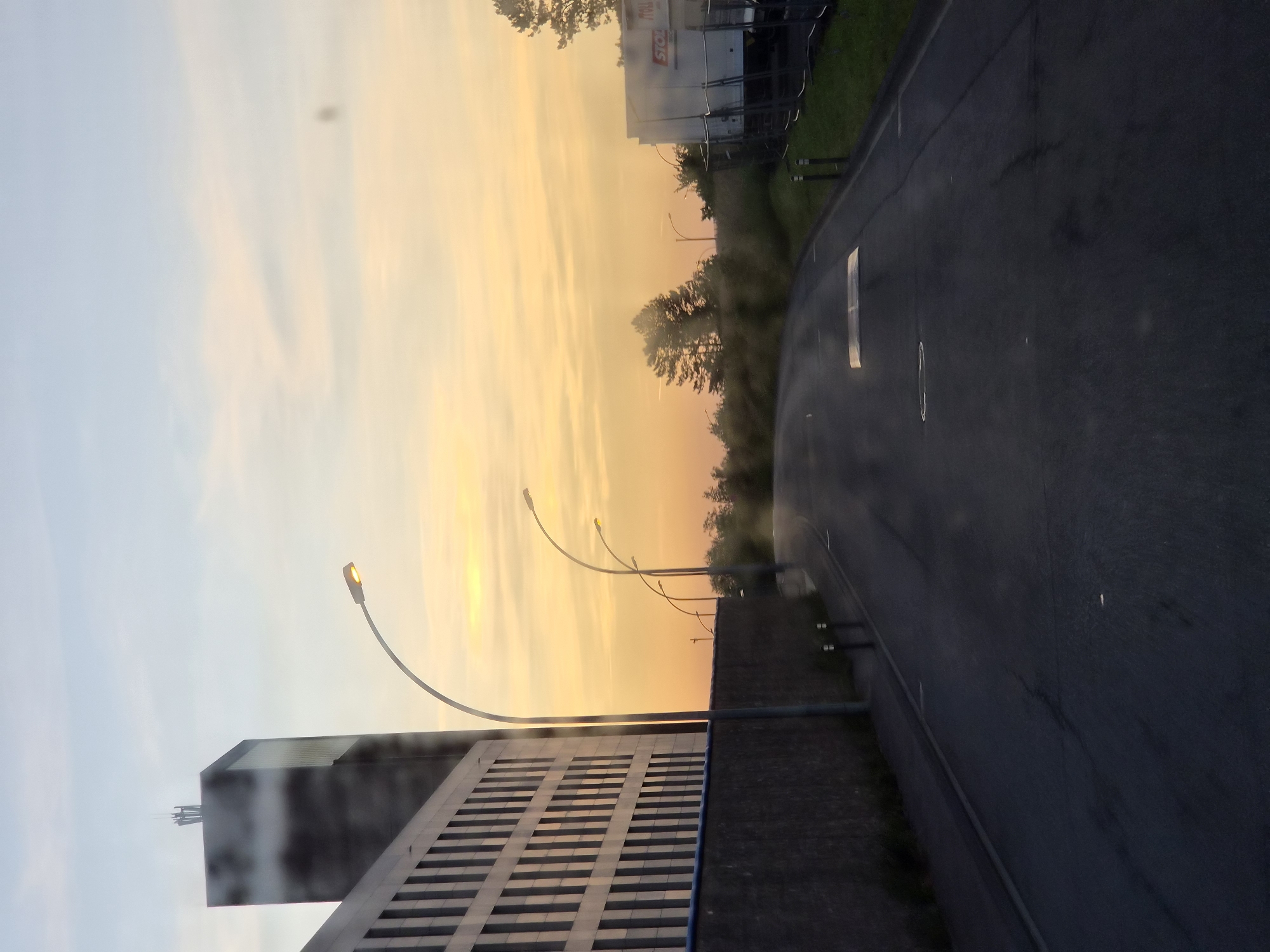 The image shows a sunset in Leudelange, Luxembourg. The sky is a mix of blue, orange, and yellow hues. A building with a reflective facade is on the left side of the image. There are street lamps along the road. A road runs alongside the building and a fence. There are trees and a white structure in the background.