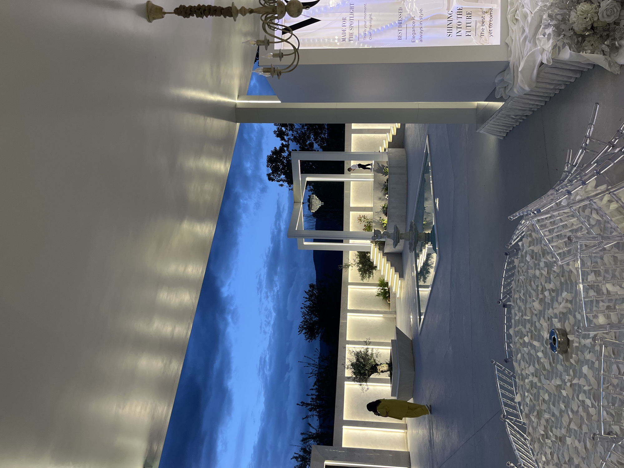 The image captures a modern architectural design possibly located in Gotse Delchev, Bulgaria, based on the presence of Cyrillic text in other images of the area. The architecture features a multi-tiered structure with recessed lighting and landscaping with a fountain. The sky is visible in the background, suggesting it is dusk or early evening. A person in a yellow outfit is walking along the lower tier. There is also a display featuring text promoting a sense of elegance and future ambition, including phrases like "MADE FOR THE SPOTLIGHT", "BEST DRESSED", and "SHINING INTO THE FUTURE". The foreground contains transparent chairs and floral arrangements, implying this could be a venue for events.