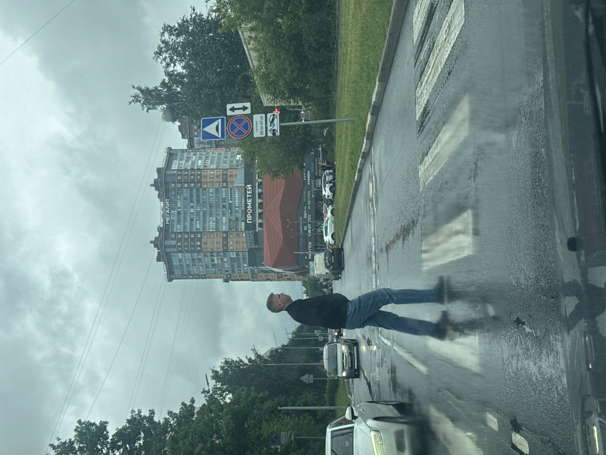 A man is crossing a wet crosswalk on a cloudy day, possibly in Saint Petersburg, Russia, given the Cyrillic text visible. He is wearing a dark jacket and blue jeans. The street is wet, indicating it has recently rained or is currently raining. The crosswalk markings are clear white stripes on the asphalt. In the background, there are several cars and a tall, modern building with many windows, bearing the Cyrillic text "МЕГАЛИТ" and "ПРОМЕТЕЙ". Traffic signs are also visible, including one with a blue triangle and a red circle with a diagonal line, indicating a prohibition. Another sign shows a red traffic light symbol and parking restrictions. The overall mood is somber due to the overcast sky and wet conditions.