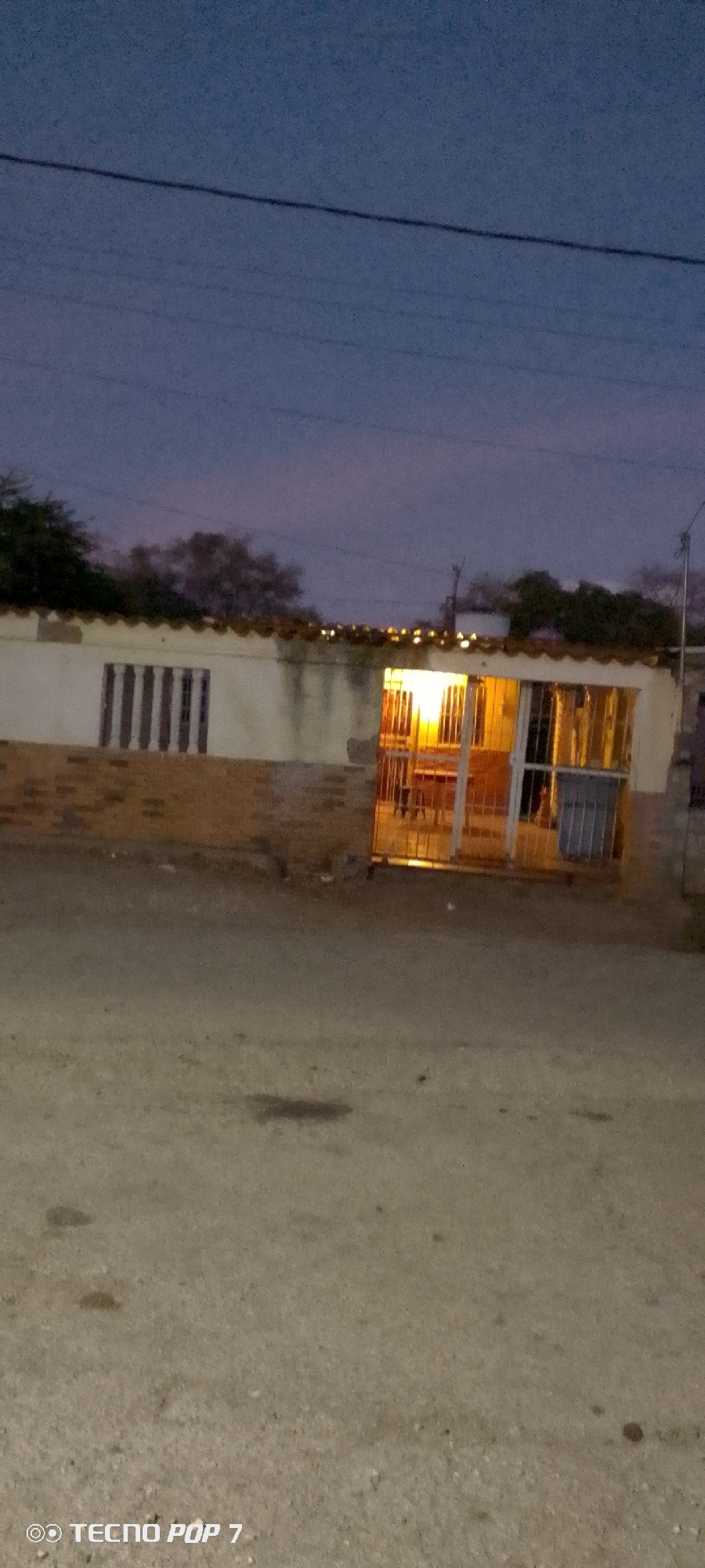 The image depicts a single-story building in Altagracia, Venezuela, at dusk. The building has a light-colored facade with a brick base and a corrugated metal or tile roof. A window with vertical bars is visible on the left side of the building. The front entrance is illuminated by an interior light, revealing a metal security gate and glimpses of furniture inside. A metal trash can is positioned near the entrance. A dirt road or street is in the foreground, with some discoloration or spots. Power lines are visible against the twilight sky. The sky is a mix of blue and pink hues. Trees are sparsely distributed in the background. The camera model is identified as a TECNO POP 7.