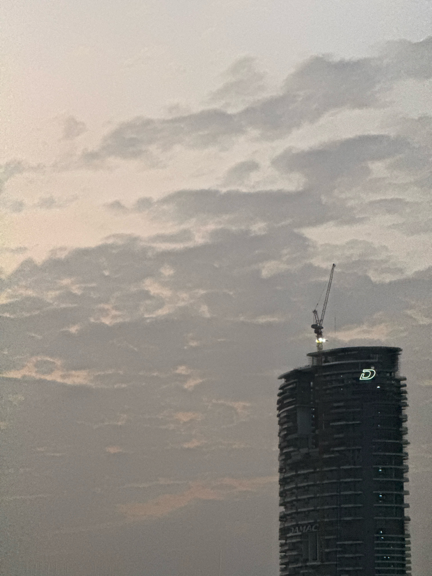 The image shows a dusk or dawn sky filled with scattered clouds over a cityscape in Dubai, United Arab Emirates. A tall, dark, modern skyscraper is prominent in the right half of the frame. The building has a curved facade with many windows and balconies. A construction crane is visible extending from the top of the building. A white illuminated logo, appearing to be a stylized letter "D", is affixed to the upper portion of the building. The name "DAMAC" is visible in smaller lettering near the base of the building. The overall scene suggests an urban environment under a diffused, overcast sky.