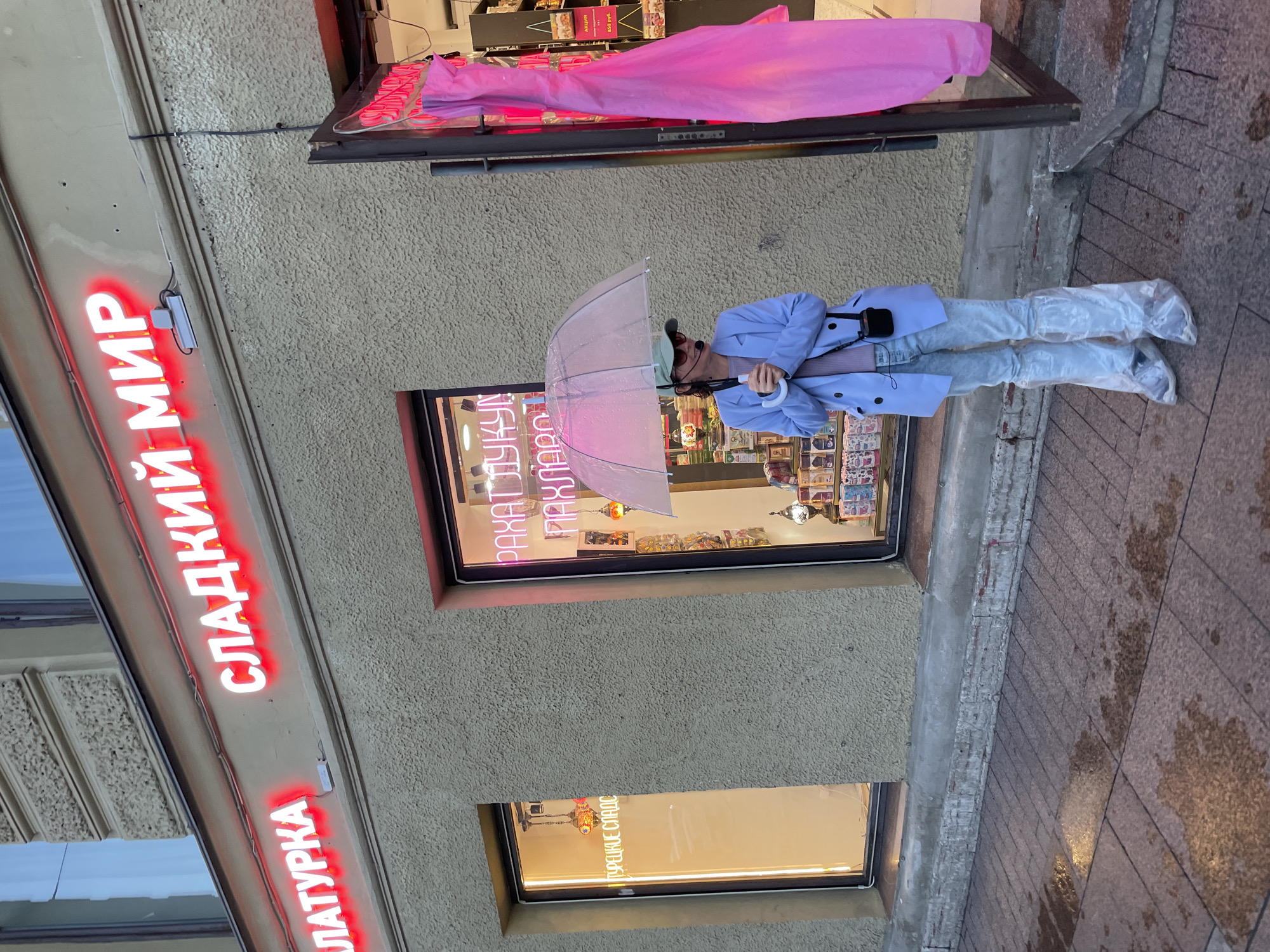 The image depicts a street view in Saint Petersburg, Russia, specifically showing the facade of a confectionery shop. The shop's name, "СЛАДКИЙ МИР" (Sweet World), is illuminated by red neon lights above its entrance. To the left of the main entrance, a window displays a neon sign that reads "ТУРЕЦКИЕ СЛАДОСТИ" (Turkish Delights). Inside the shop, various goods are visible, including shelves stocked with packaged items and decorative lamps.
A woman is standing on the wet sidewalk in front of the shop, holding a transparent umbrella. She is wearing a light blue jacket over a light purple shirt, light-wash jeans, and clear plastic shoe covers over her sneakers. She appears to be looking to her right, away from the camera. A small black device, possibly a microphone or camera, is attached to her jacket.
The sidewalk is paved with reddish-brown tiles and shows signs of recent rain with puddles reflecting the light from the shop. A large, bright pink fabric hangs vertically outside the shop entrance to the right. The building's facade is textured and painted in a beige color. The overall scene suggests an overcast or rainy day.