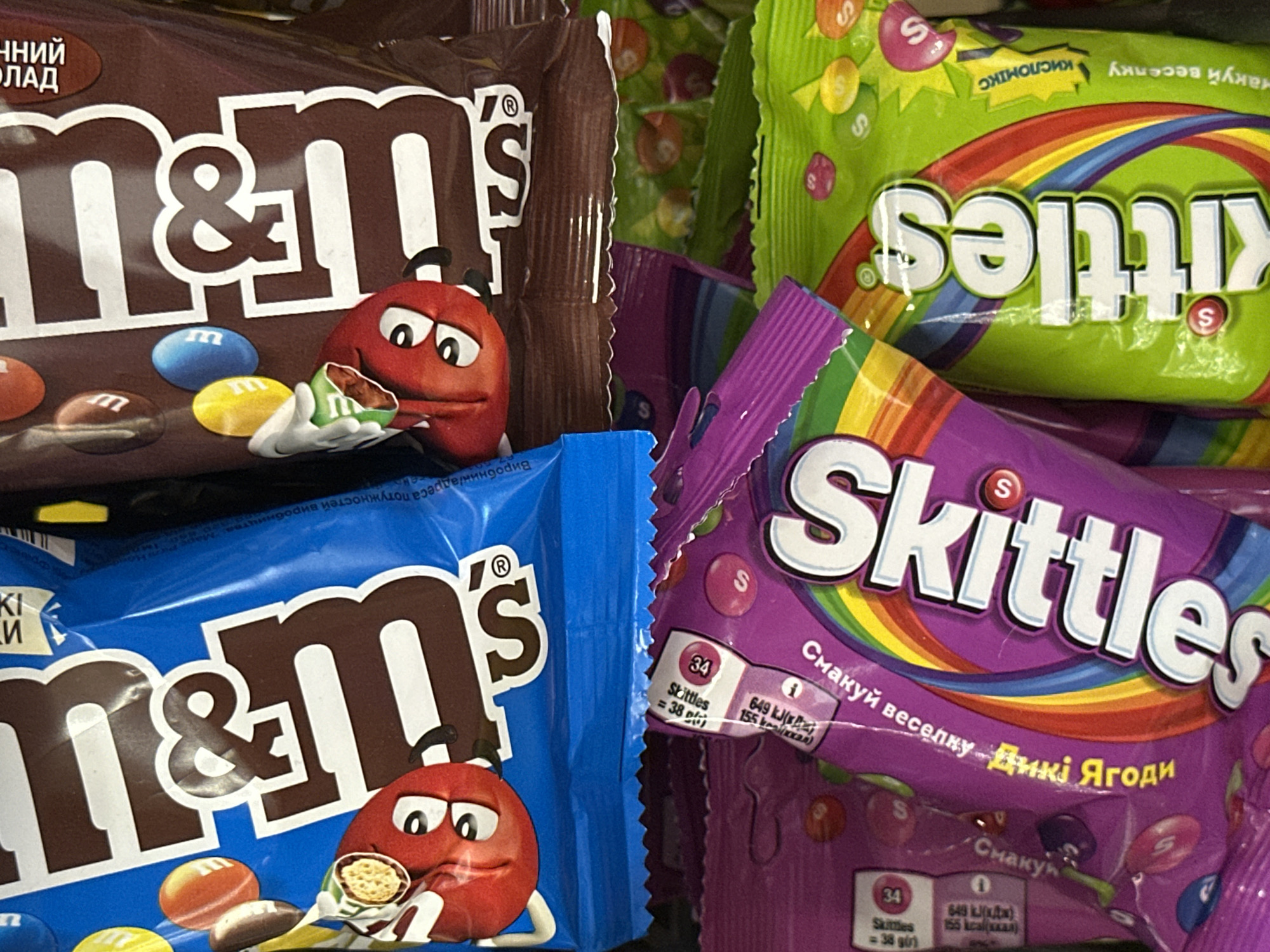 The image displays a close-up, overhead view of several packages of M&M's and Skittles candies, likely in a retail setting. The candies are arranged in a somewhat jumbled manner, with the bright colors of the packaging dominating the frame.
In the upper portion of the image, two large packages of M&M's are visible. The left package is blue and prominently features the M&M's logo in large white and brown letters. A cartoonish red M&M character with big eyes is visible on the blue packaging. To the right, a brown package of M&M's is partially visible, also displaying the brand's logo.
Below the M&M's, two packages of Skittles candies are present. The left package is purple and features the "Skittles" logo in large white letters with a rainbow motif. Text in what appears to be Cyrillic script is visible on this package, along with nutritional information labels. The right package is bright green and also shows the "Skittles" logo and rainbow design. Similar Cyrillic text and rainbow elements are visible on this package as well. Several individual candies, colored red, yellow, and purple, are scattered amongst the packaging.
The lighting in the image is bright, suggesting it was taken under artificial store lighting or with a flash. The focus is sharp on the candy packages, with a shallow depth of field rendering the background slightly blurred. No people or distinct environmental features are identifiable, indicating the focus is solely on the confectionery products.