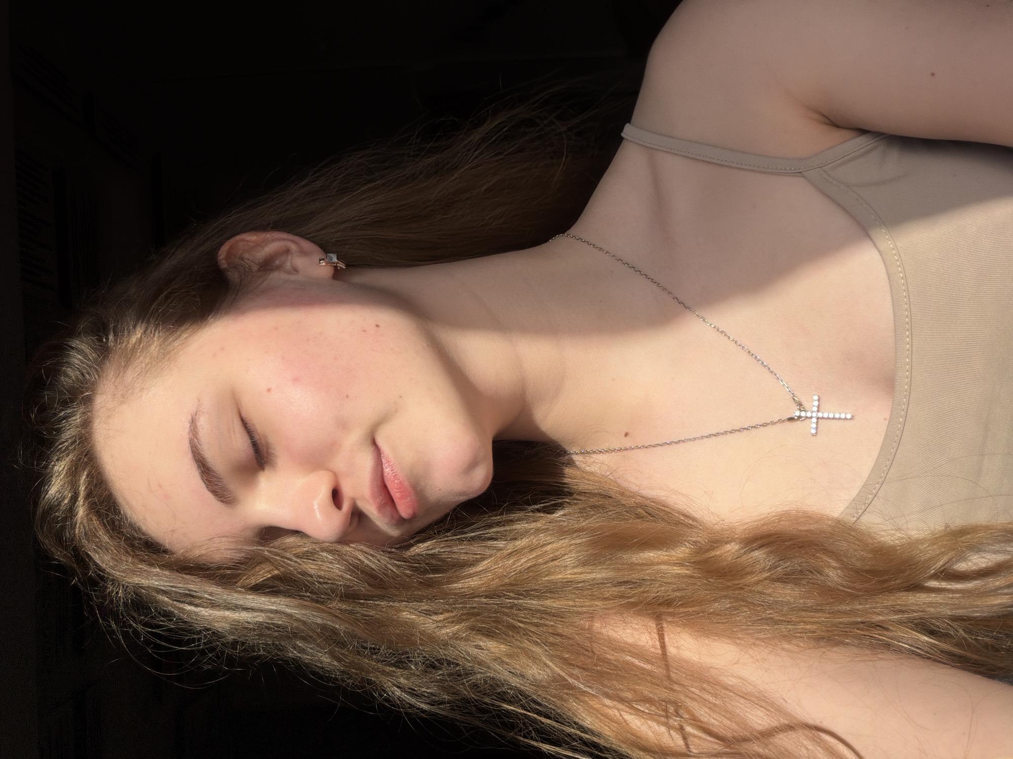 The image shows a close-up of a young woman's face and upper body. She has long, wavy, light brown hair that falls over her left shoulder. Her eyes are closed, and her lips are slightly parted. She is wearing a delicate silver necklace with a cross pendant adorned with small white stones. She also has a small silver earring in her right ear. A shadow falls diagonally across her face and chest, creating a stark contrast with the sunlight. She is wearing a beige, spaghetti-strap tank top. The background is dark and out of focus, suggesting an indoor setting. There is no identifiable information regarding the location in Dnipro, Ukraine.
