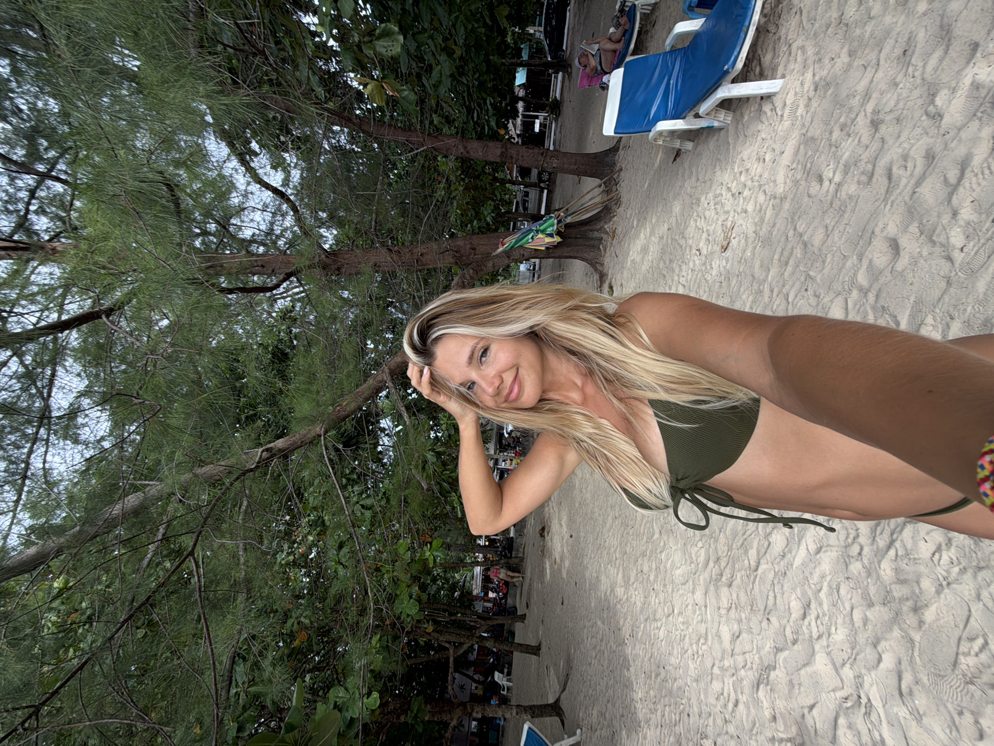 A woman with blonde hair, wearing a green bikini, stands on a sandy beach in Sakhu, Thailand, holding her phone to take a selfie. Trees with lush green foliage line the background, with several beach chairs and umbrellas visible. The sand is light-colored and textured with footprints. In the distance, several people are seen near a row of buildings, suggesting a beachside village or resort. The sky is overcast.