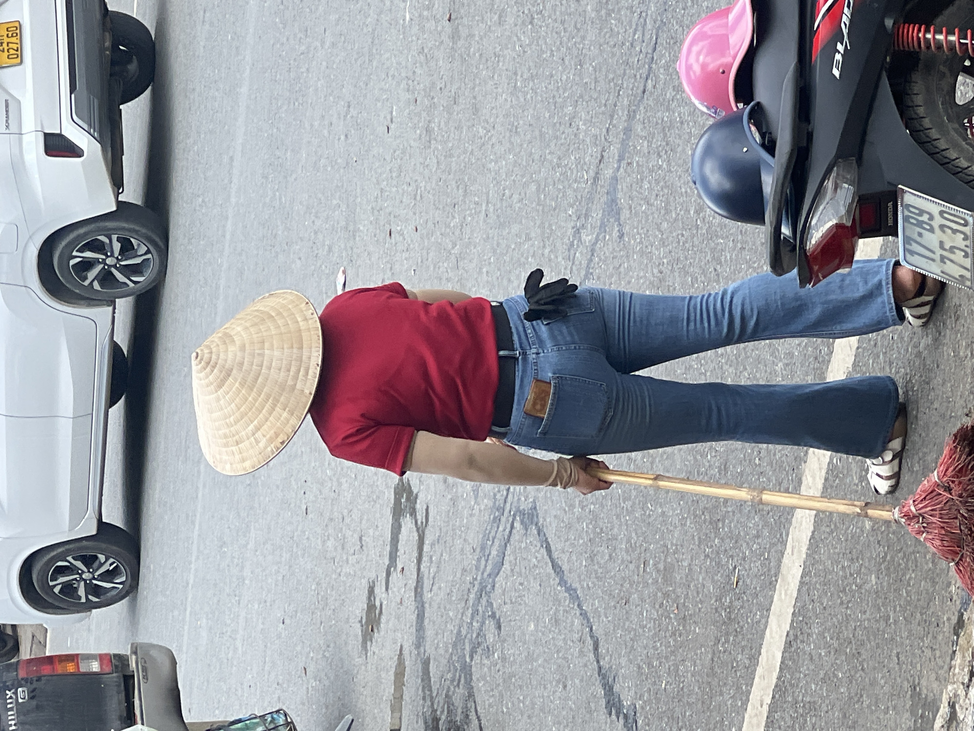 In Sa Pa Ward, Vietnam, a person wearing a conical hat, a red t-shirt, and blue jeans stands on a paved street, holding a broom with a bamboo handle and a red brush. The person is seen from the back, with a black glove tucked into their right back pocket and a wallet in their left back pocket. Behind them, a white car is parked. In the foreground, the back of a black motorcycle is visible, with a dark blue helmet and a pink helmet placed on the seat. The ground is asphalt with white markings.