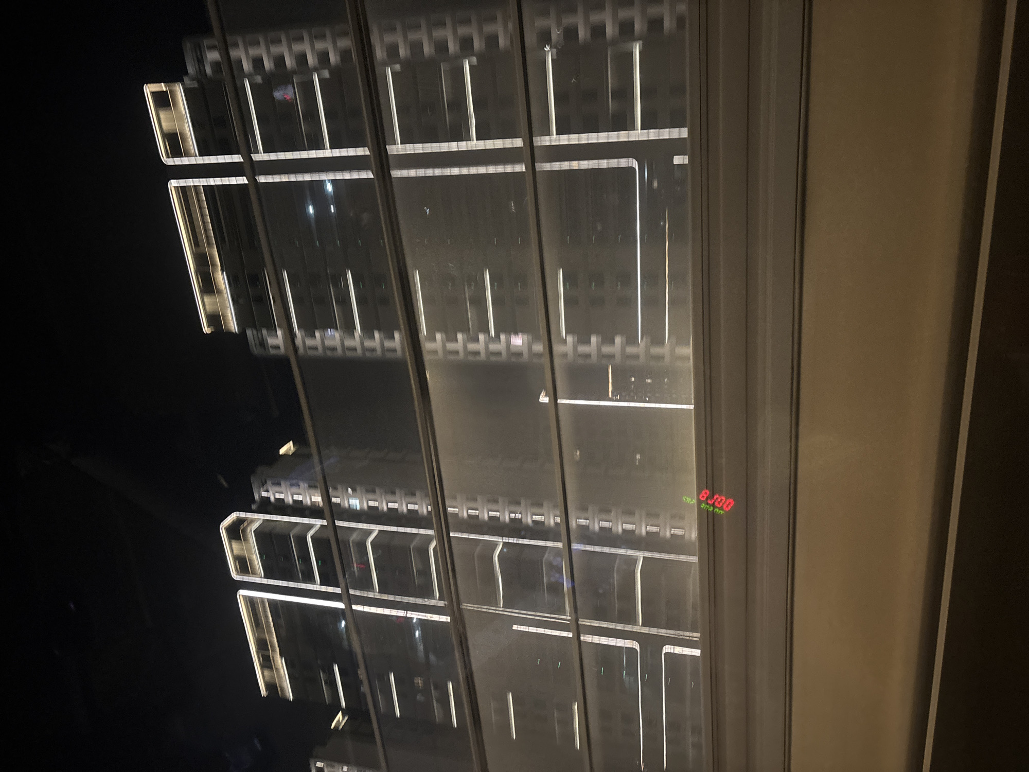 The image captures a night view of modern high-rise buildings in Xiangcheng District, China, as seen through a window or a transparent barrier. The architecture is characterized by sleek lines and extensive illumination, with horizontal and vertical LED strips accentuating the building facades. Two prominent towers dominate the scene, their upper sections topped with warm-toned lights, suggesting a contemporary urban landscape. The buildings appear to be residential or office complexes, with multiple stories and visible windows. Reflections on the transparent surface create a layered visual effect, adding depth to the composition. In the foreground, a horizontal barrier or railing is present, with a small, illuminated red digital display showing what appears to be "8.00" at the lower left. The overall atmosphere is nocturnal and somewhat hazy, possibly due to atmospheric conditions or the quality of the glass. The background is dark, indicating a nighttime setting in a city environment.
