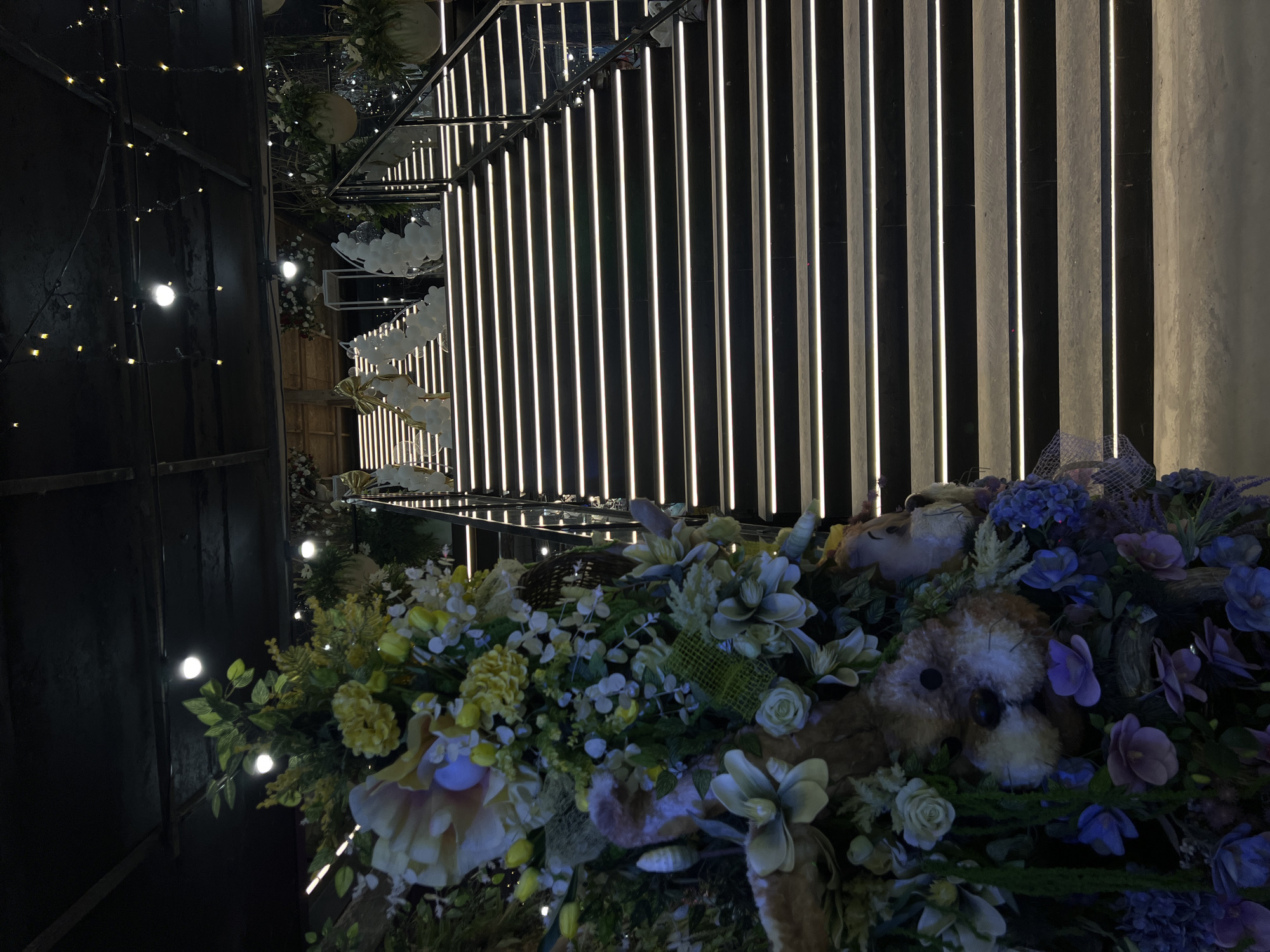 The image depicts a grand, illuminated staircase with plush animal figures and floral arrangements in the foreground, suggesting a decorative event setting. The staircase is wide and appears to be made of dark stone, with each step outlined by a bright, horizontal LED light strip. The top of the staircase leads to a doorway or entrance, adorned with white balloons and golden bows.
In the foreground, to the left, a large, elaborate floral arrangement is visible, incorporating plush animal toys, flowers in shades of yellow, white, and purple, and greenery. These elements create a whimsical and festive atmosphere.
The background shows a dark ceiling with exposed beams and hanging lights, contributing to the overall ambiance. The scene suggests an indoor venue, potentially a banquet hall or a reception area, decorated for a special occasion. The style and decoration do not provide specific clues to its location within Ternopil, Ukraine, or any other city. The image is devoid of any people or discernible actions.