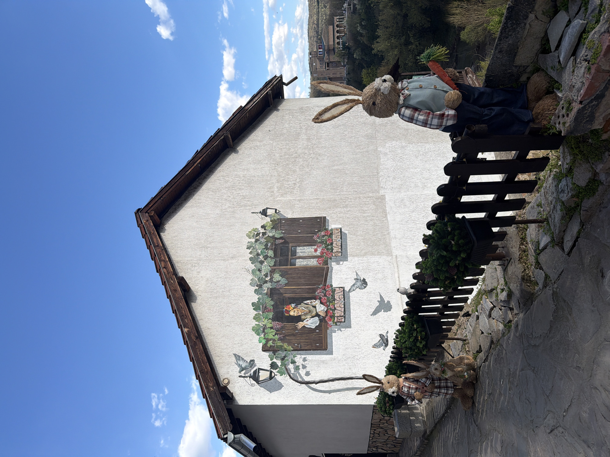 A building with a mural on its wall stands under a blue sky with scattered clouds. The mural depicts a woman looking out of a window with flowers and vines around it and pigeons are also present. There are two stuffed rabbits dressed in plaid shirts present, one sitting on a fence holding a carrot. Veliko Tarnovo, Bulgaria can be seen in the background. The fence and wall are stone.