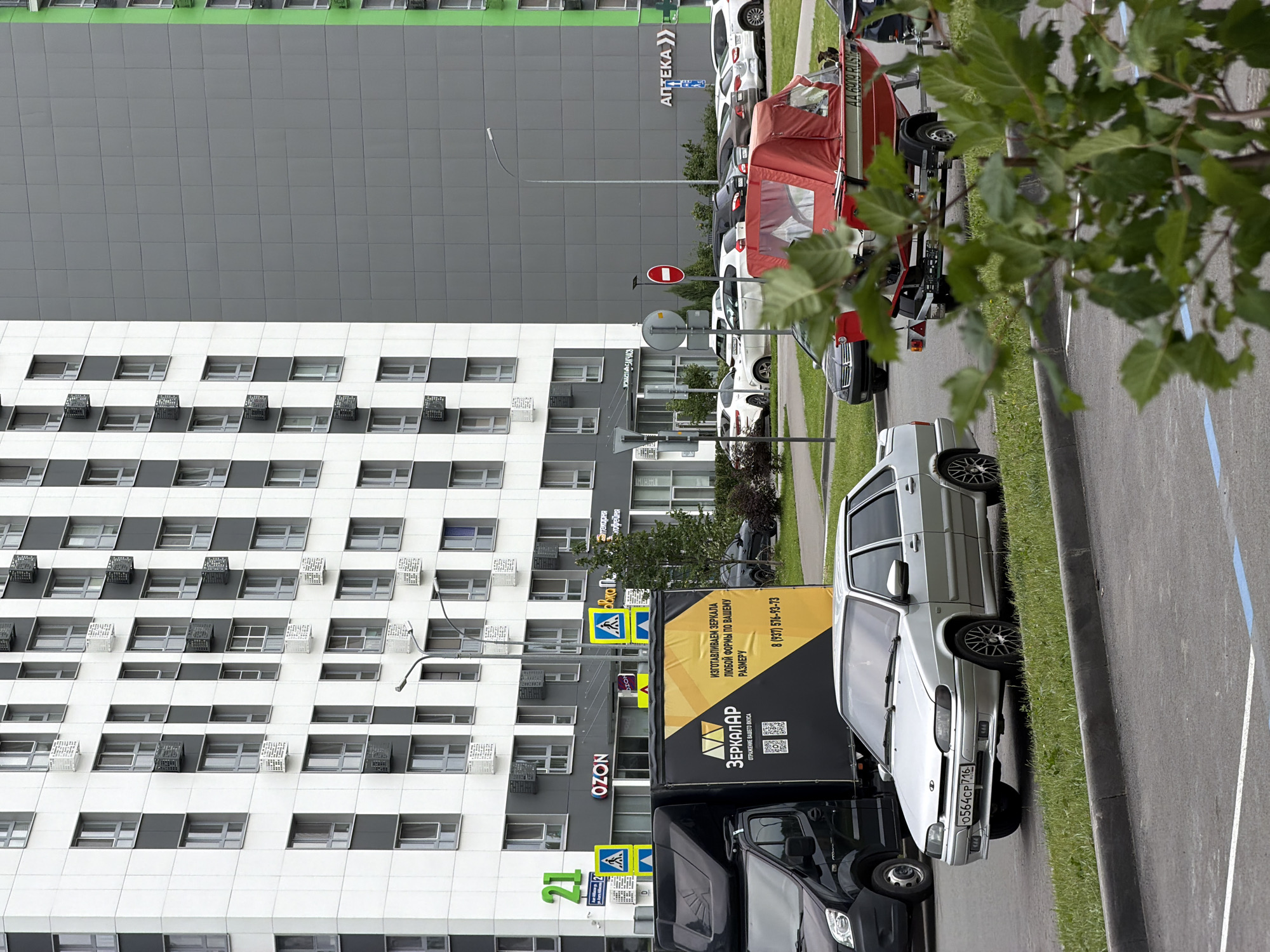 This media file depicts a street scene in Kazan, Russia, featuring a modern, multi-story building with a predominantly gray and white facade. Numerous windows and what appear to be air conditioning units are visible on the building's exterior.
In front of the building, a row of parked vehicles is lined up along the roadside. These include a silver sedan, a black truck with a large yellow and black advertisement on its side, and other cars partially visible. The advertisement on the truck prominently features the word "ЗЕРКАЛАР" (Zerkalar) and contact information.
Several street signs are also visible, including directional signs and a red circular sign with a white horizontal bar, which typically indicates a prohibition. The area is lined with a grassy verge and a paved road, suggesting an urban or suburban environment.
The presence of trees with green leaves indicates it is likely daytime and warm weather. The overall scene appears to be a typical street view in a modern Russian city, possibly a commercial or residential area. The image is oriented vertically, with the building occupying the left portion and the street and parked cars on the right. No people are clearly discernible in the image. The visible text includes business names like "OZON," "Аптека" (Apteka - Pharmacy), and the advertisement on the truck.
