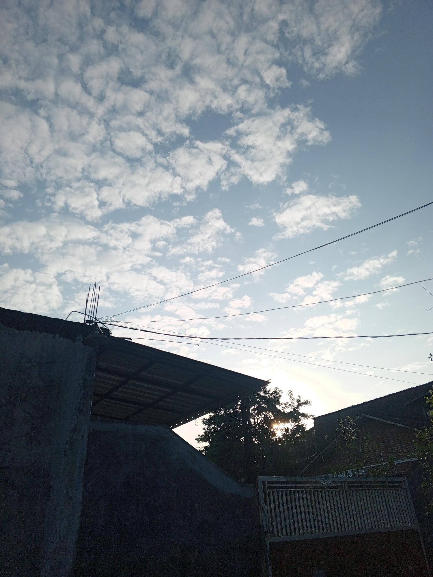 The image depicts a clear sky with scattered clouds, possibly taken at dawn or dusk, as indicated by the gentle sunlight filtering through trees and structures. The scene is set in a residential area, with the roofs of houses and a weathered concrete wall in the foreground. Electrical wires are strung across the sky, a common sight in urban and semi-urban environments. The overall impression is one of a peaceful, everyday moment in a neighborhood. The location is identified as Sumbersari, Indonesia, suggesting a tropical or subtropical climate given the lushness of the trees visible.