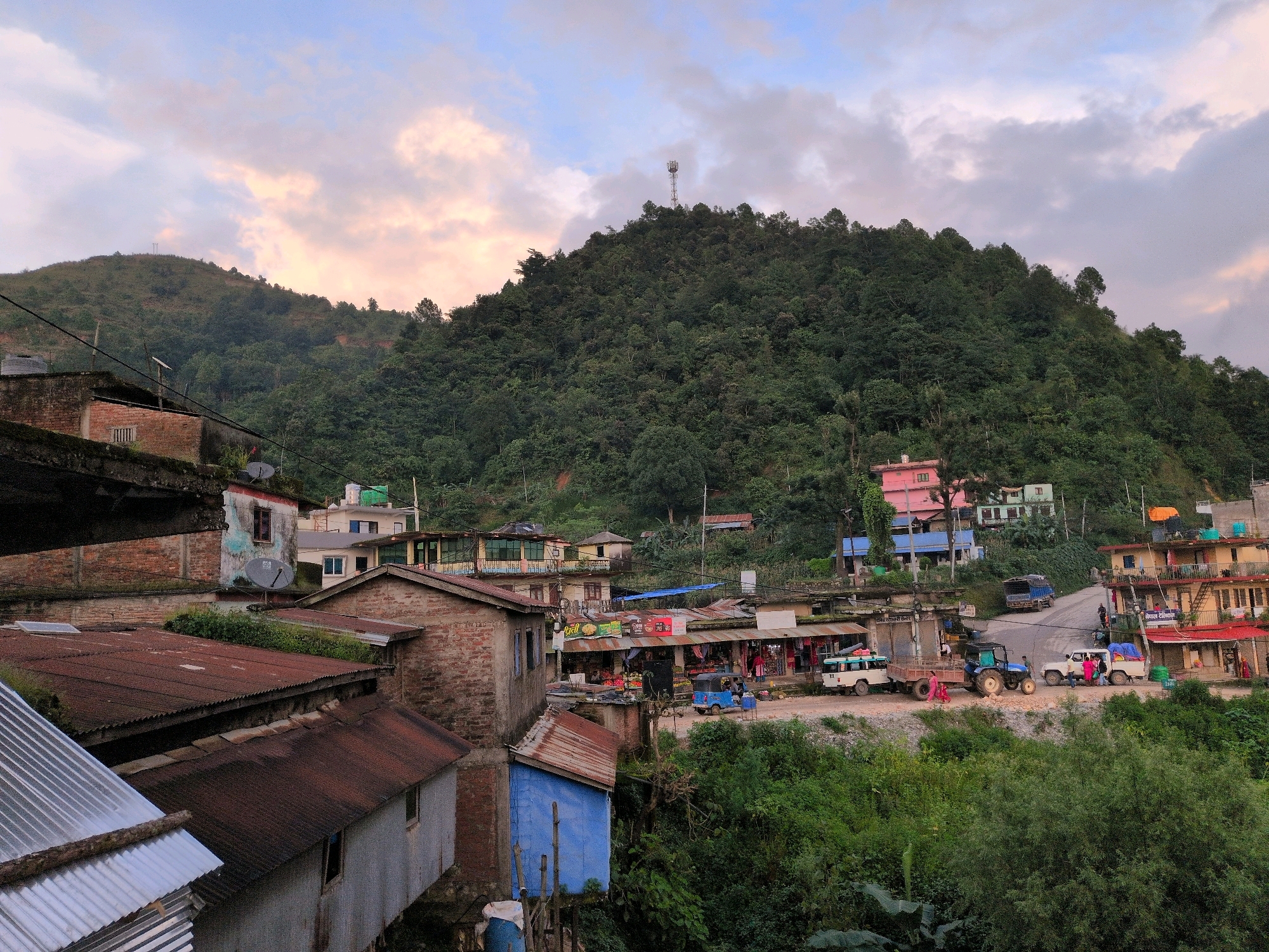 The image depicts a hillside village scene in Kapurkot, Nepal, under a cloudy, pastel sky. A dense, green forest covers the upper slopes of a large hill, capped by a cell tower. In the foreground and mid-ground, a cluster of buildings, many with corrugated metal roofs, are nestled into the hillside. The architecture appears rustic, with a mix of brick and painted structures. A paved road winds through the village, with several vehicles, including a blue tractor, a white SUV, and a white minivan, present. People are visible near the vehicles and in front of small shops lining the road. Lush vegetation surrounds the buildings, indicating a natural and somewhat untamed environment. The overall impression is of a remote, developing community situated within a mountainous landscape.