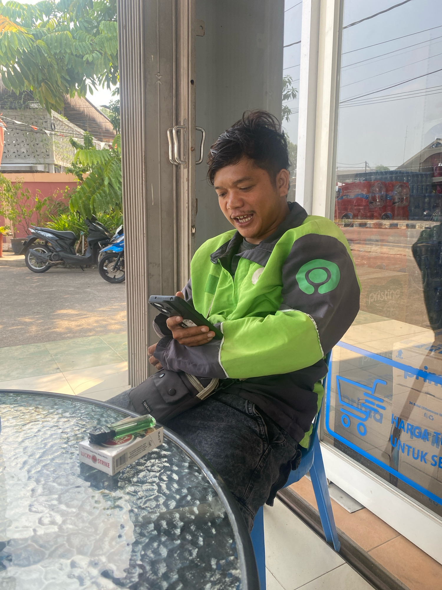 A man wearing a green and gray jacket with a green circular logo sits at a glass-topped table, looking at his phone. On the table in front of him is a pack of Lucky Strike cigarettes and a green lighter. The man is sitting on a blue chair outside a building with a large, closed metal gate to his left and a window to his right. Through the window, a street with parked motorcycles and a red truck can be seen. The environment suggests an outdoor seating area of a business in the city of Tanjung Redeb, Indonesia, likely a cafe or shop.