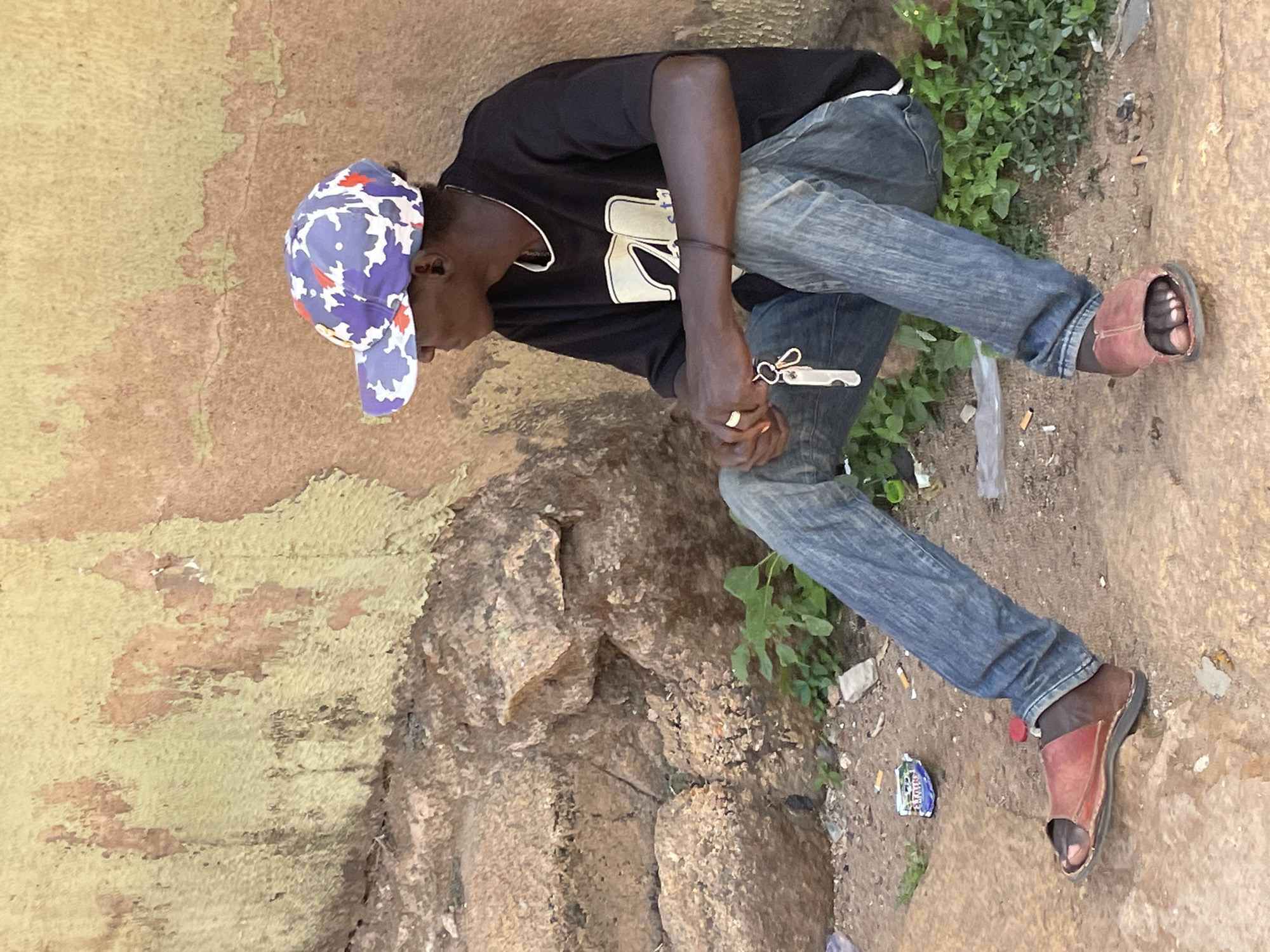 A Black male wearing a black t-shirt with gold decals, faded blue jeans, and burgundy sandals sits on a rock outcropping. He wears a blue and purple patterned cap. He faces left. The background consists of a yellow-painted wall with decaying paint, the rock formation on which he is seated, and ground with weeds. A package of chewing gum and cigarette butts lay on the ground near his left leg. The location is likely Jos, Nigeria.