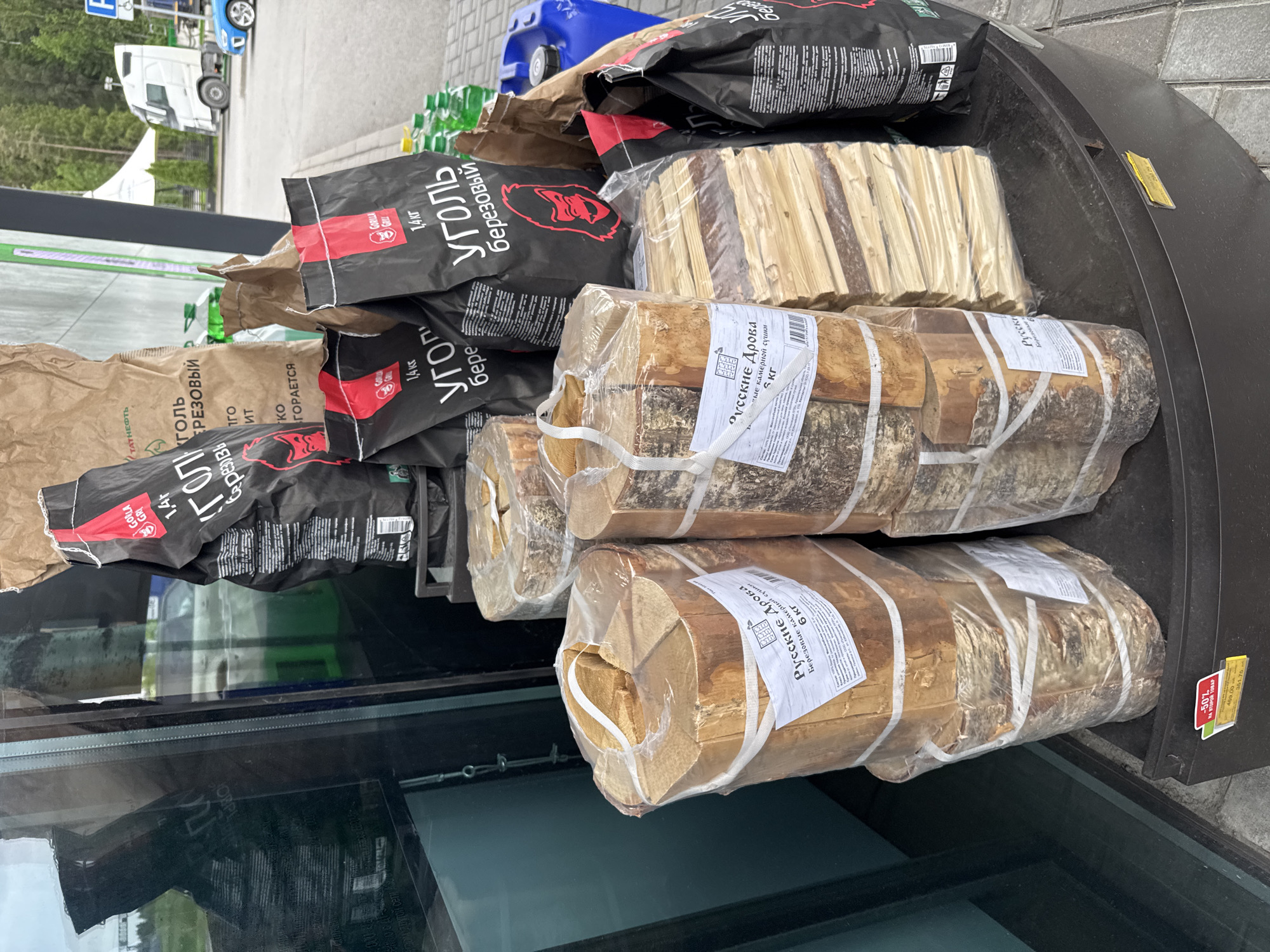 The image shows packages of firewood and charcoal displayed on a black, rounded surface outside a building, likely a store, in Medovoye, Russia. The firewood packages are wrapped in clear plastic with white labels in Russian, indicating "Russian Firewood, Birch, kiln-dried, 6 kg." Bags of charcoal, some labeled "Gorilla Grill 1.4 kg", are stacked behind the firewood. A small, yellow price tag is visible on the right edge of the display. The reflections in a glass surface indicate an outdoor environment, with a truck and trees visible in the background.