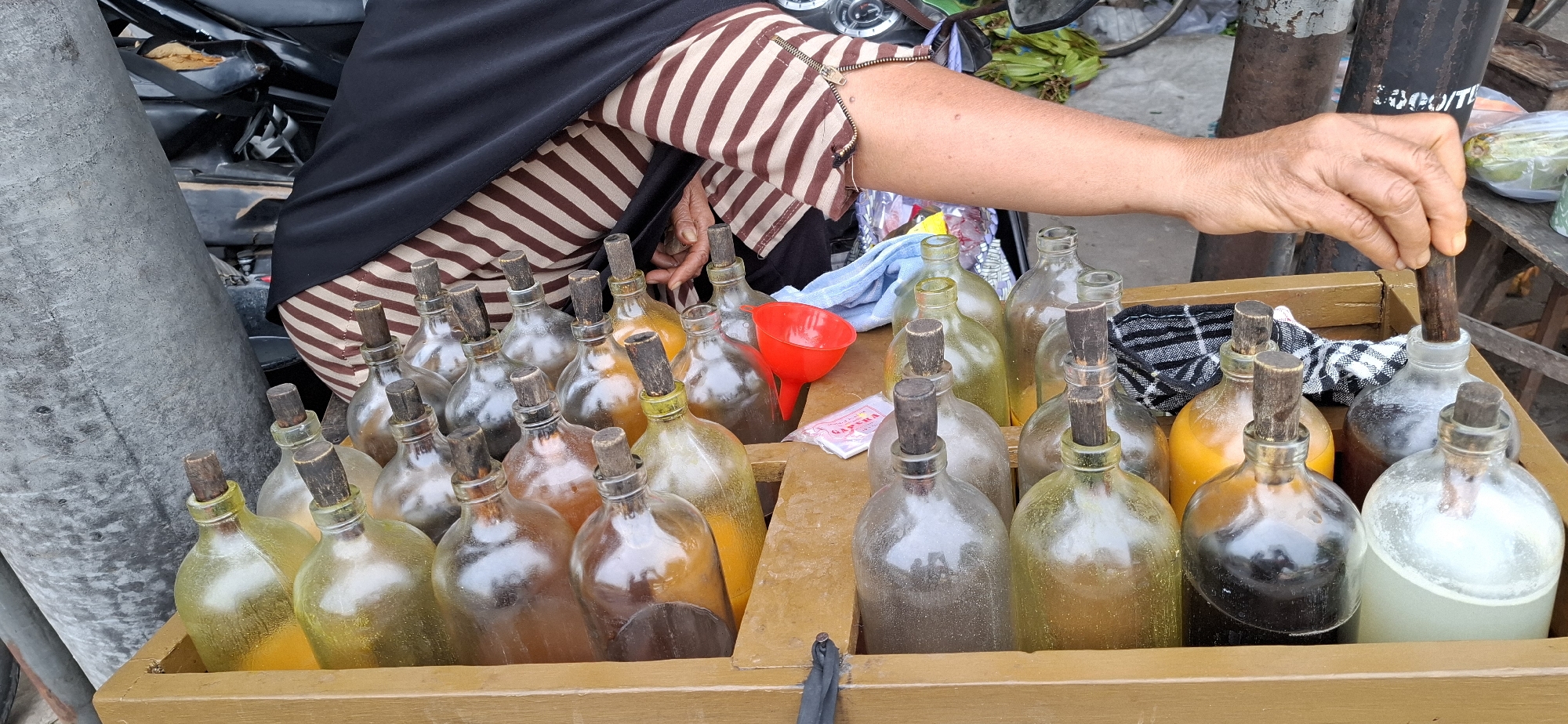 In Mranggen, Indonesia, a vendor displays numerous glass bottles filled with various colored liquids, likely traditional Indonesian herbal remedies or traditional fuel. The bottles, with wooden stoppers, are arranged in a wooden crate. A person, partially visible from the torso down, is wearing a striped brown and white garment and a black headscarf. Their right hand is actively engaged, holding a wooden stick and inserting it into one of the bottles, suggesting they are either measuring or dispensing the contents. A small red funnel and a packet of what appears to be a product label are also present on the crate, indicating a commercial transaction is underway. The background hints at an outdoor market setting, with a motorcycle and other market elements partially visible. The overall scene depicts a typical street-side vendor in Mranggen, Indonesia.