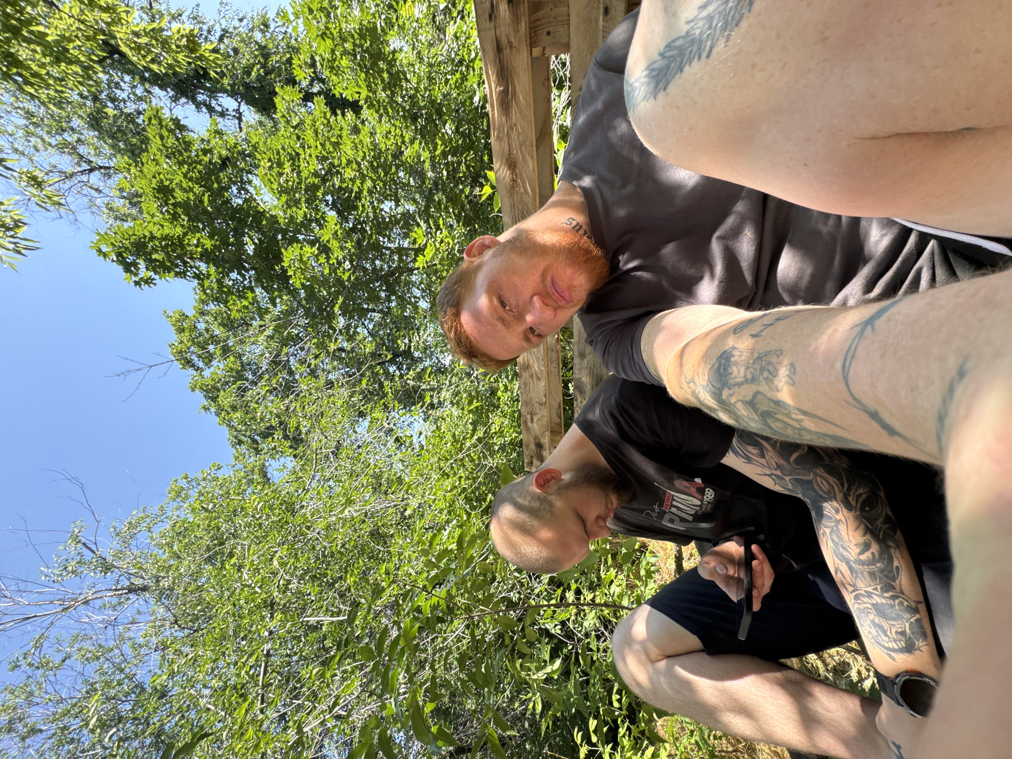 Two men are sitting on a wooden bench outdoors. The man on the left, with a shaved head and beard, is looking down at his phone. He is wearing a black t-shirt and dark shorts. He has extensive tattoos on his left arm. The man on the right has red hair and a beard, and he is looking towards the camera with a grimace. He is wearing a dark t-shirt and has tattoos on his neck and right leg. The background is filled with lush green trees and a clear blue sky. The lighting suggests it is a sunny day.