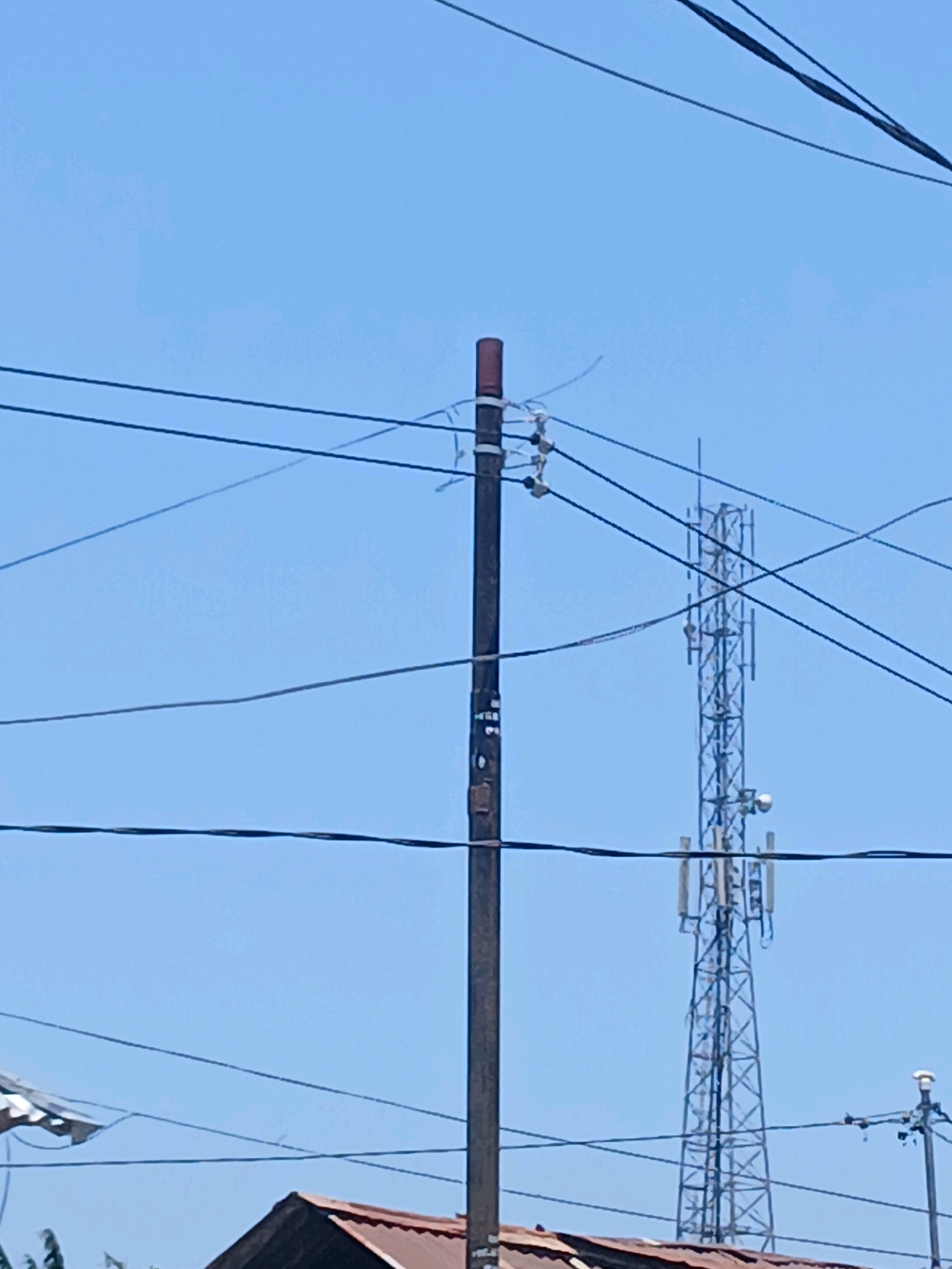 A close-up, slightly blurry, upward-angled shot shows a clear blue sky behind a dark brown utility pole with several black wires strung across it. The pole has a reddish-brown cap at the top and some metallic fixtures near the top. Further in the background, a tall, lattice-style cell tower with multiple antennas is visible. To the left and bottom of the frame, the top of a brown-tiled roof is visible, suggesting a residential area. This scene appears to be from a street in Gorontalo, Indonesia, characterized by its clear skies and the presence of essential infrastructure like power lines and communication towers.