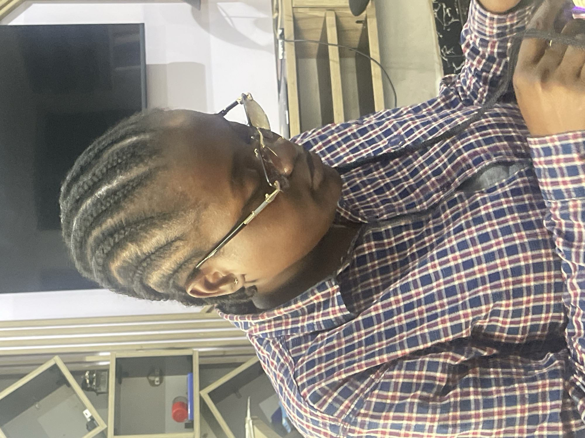 A medium shot captures a person with braided hair and glasses, looking down and focusing intently on something out of frame, likely an object or task. The individual is wearing a blue and red plaid shirt. The background includes a shelf unit with various items, a television screen, and a desk with drawers, suggesting an indoor environment that could be an office or a living space in Abuja, Nigeria. The lighting appears to be artificial, creating soft shadows and highlighting the subject. The overall scene conveys a sense of concentration and engagement with an activity.