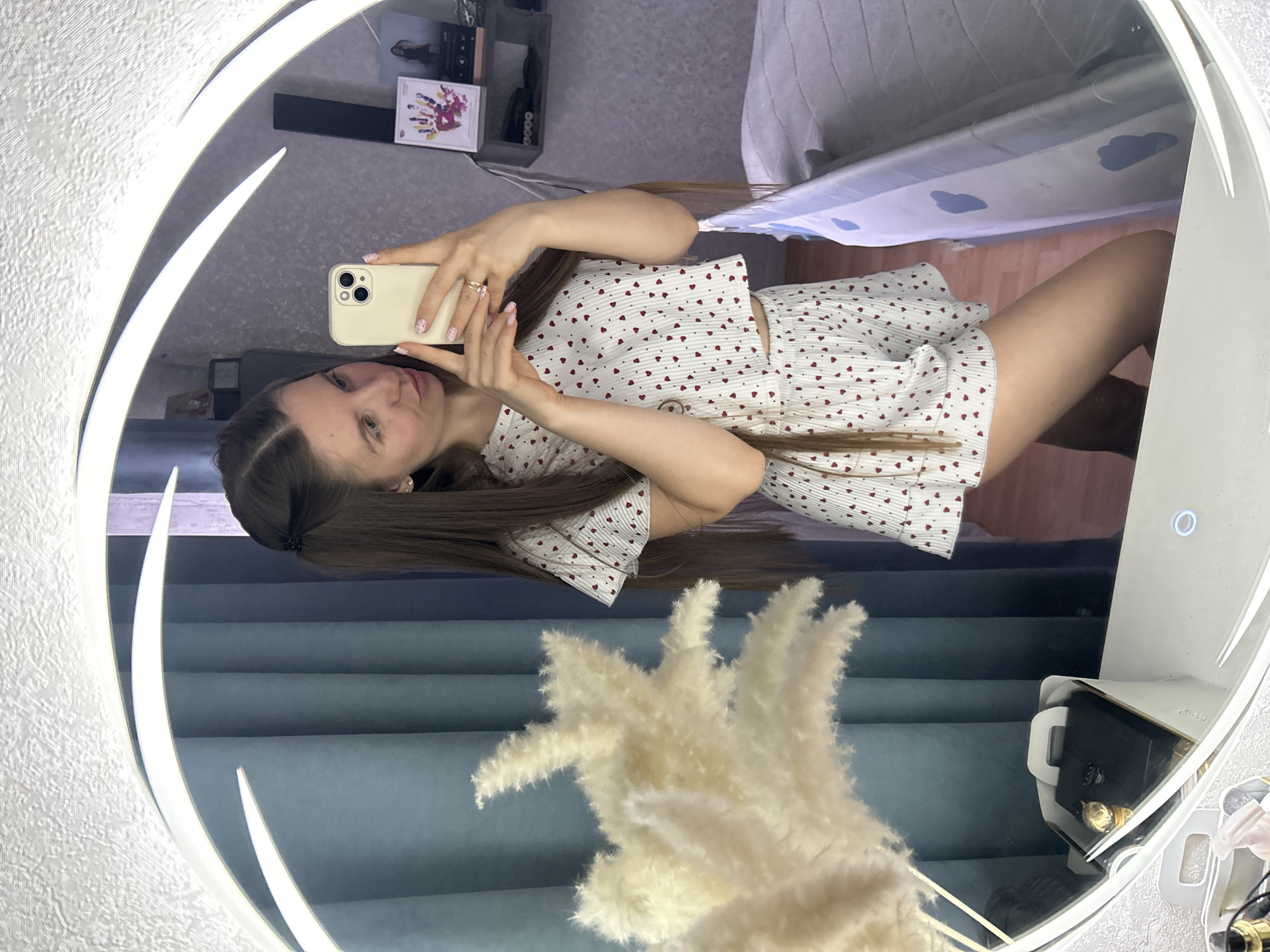 The image shows a reflection of a woman taking a selfie in a circular mirror with a light ring. The woman is holding a phone in her right hand. She is wearing a white outfit with red hearts, consisting of a short-sleeved top and shorts. She has long brown hair. The reflection shows part of a room: a bed with a blue and white patterned sheet, a shelf with various items, and a blue textured wall with a bouquet of beige pampas grass in front of it. An open jewelry box is visible. The location appears to be an interior setting. There is no evidence of the city of Klin, Russia in the image.